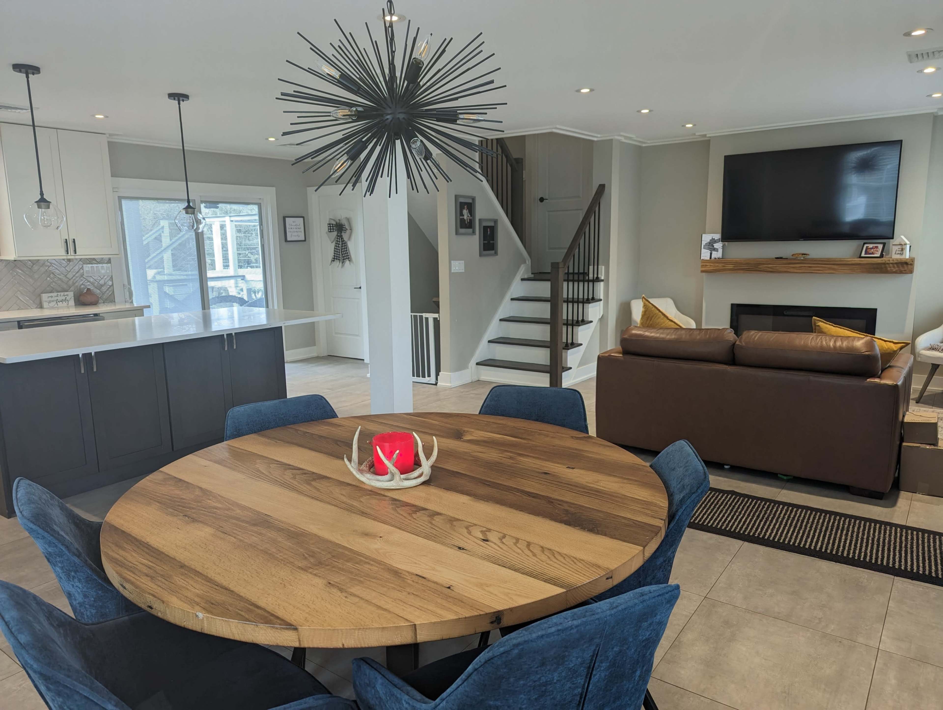 A modern living space featuring a round wooden dining table, blue upholstered chairs, a brown couch, and a staircase leading to an upper level, with a decorative light fixture and a television mounted on the wall.