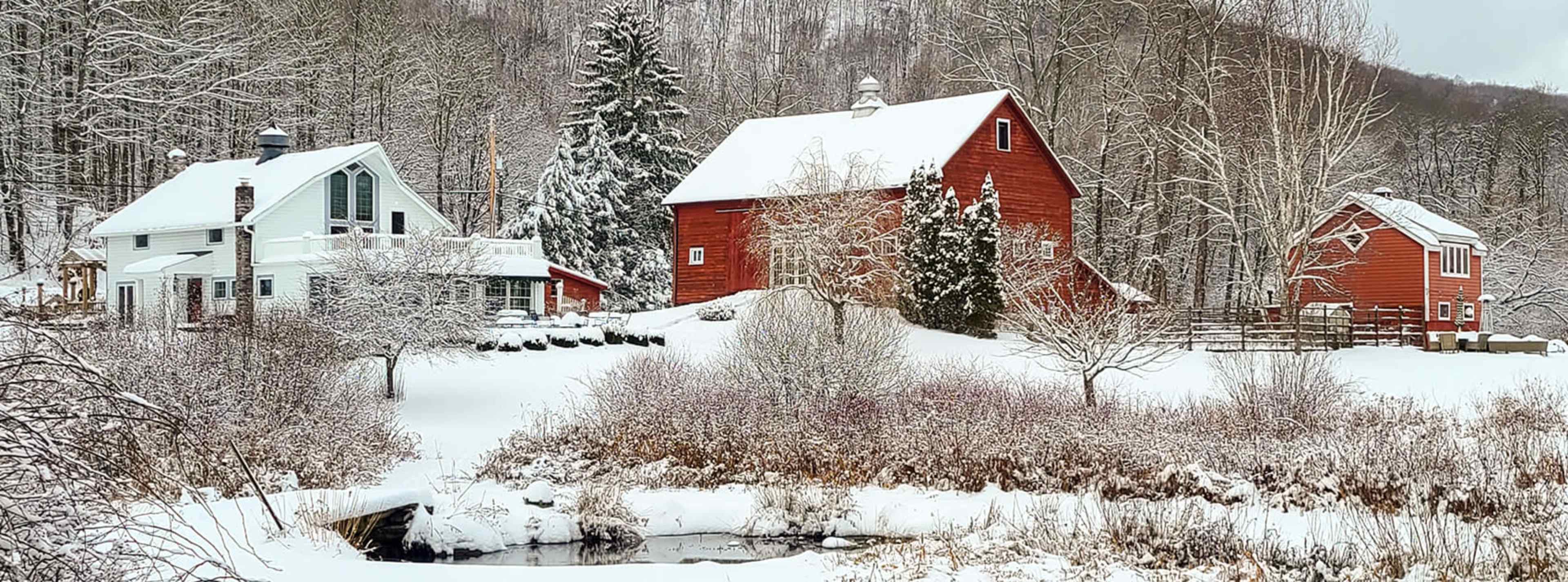 A red barn and two houses are surrounded by snow-covered ground and trees in a winter landscape.