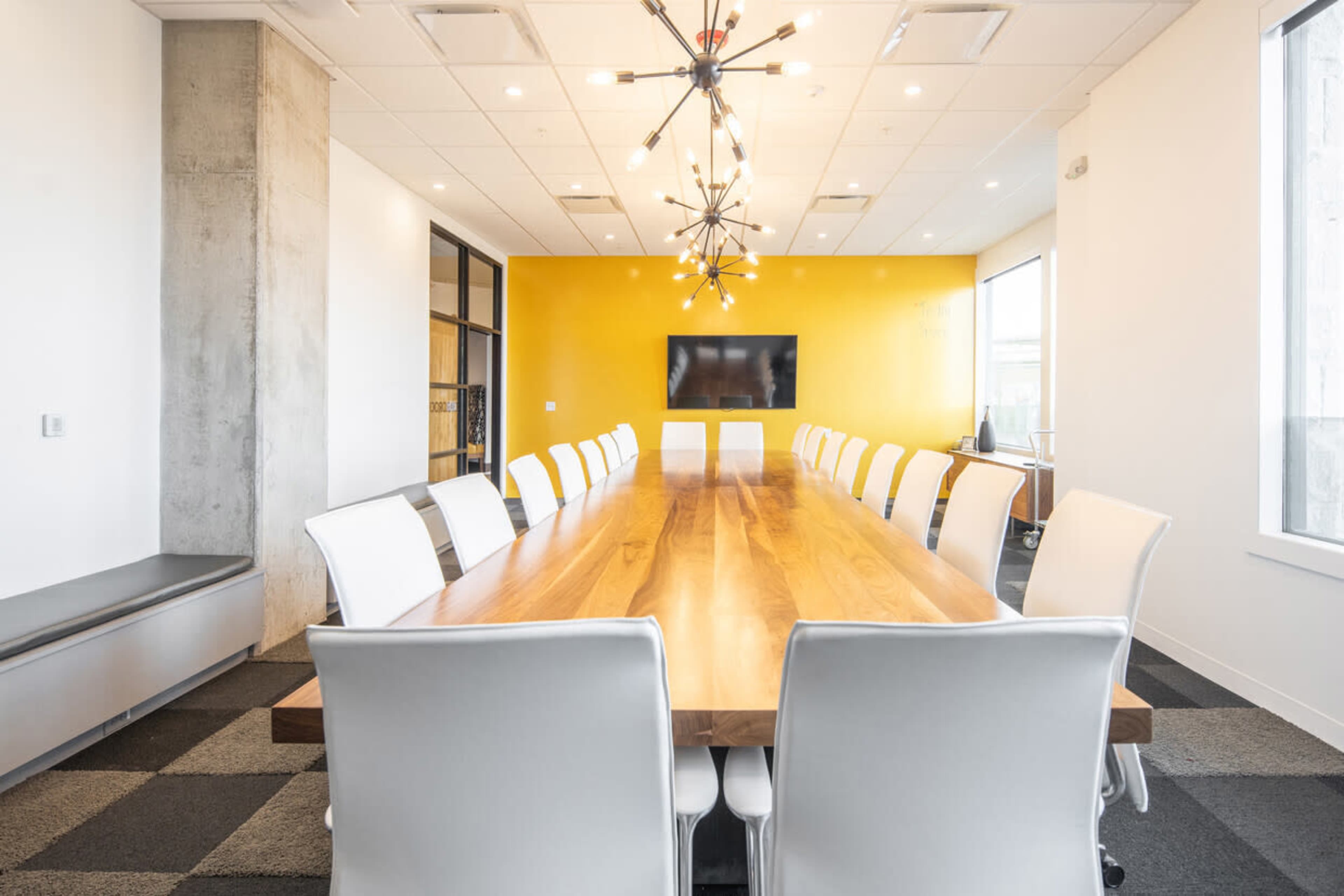 A long wooden conference table with white chairs is set in a brightly lit meeting room featuring a yellow accent wall and a modern light fixture.