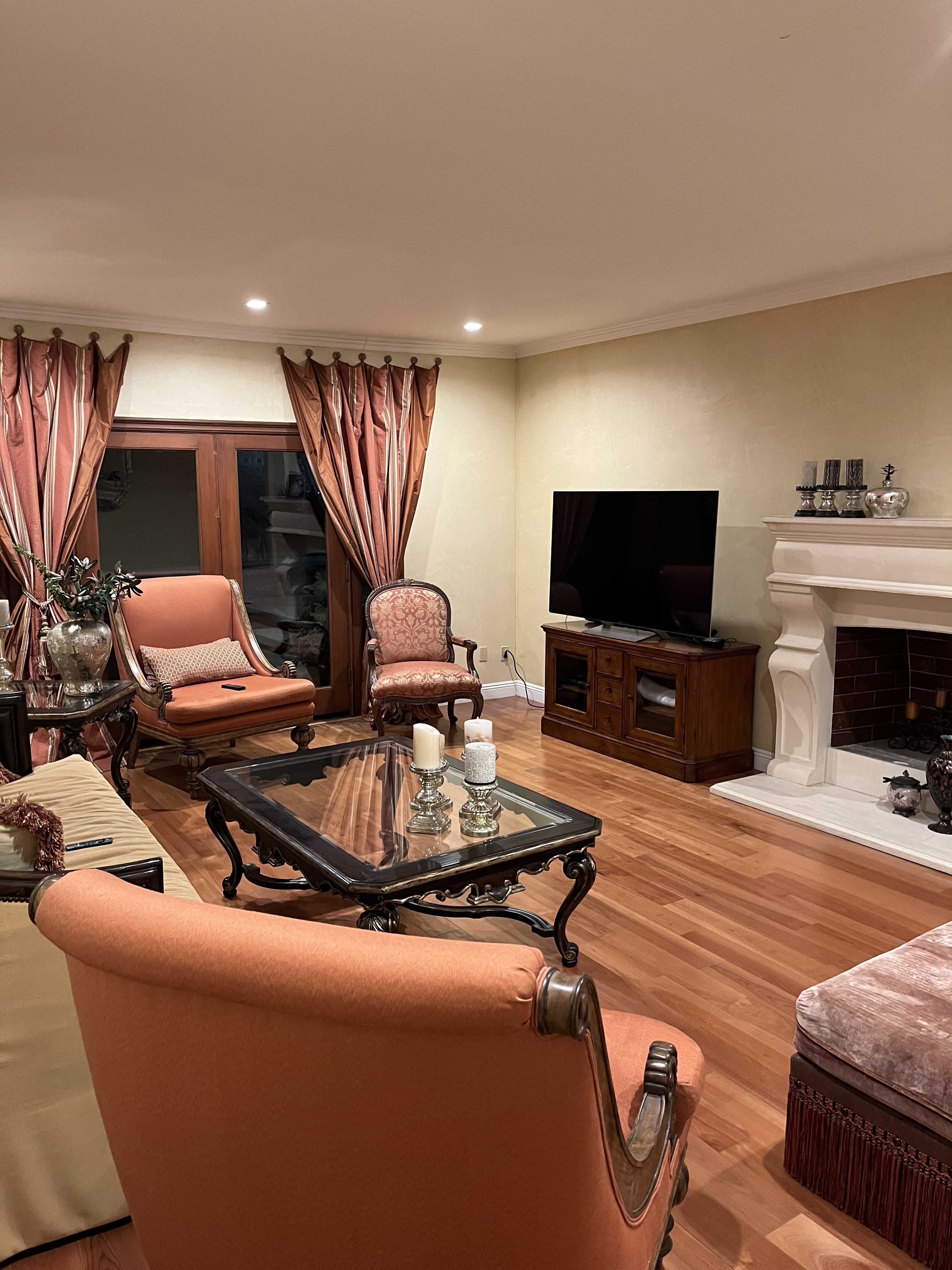 A spacious living room with wooden flooring, elegant curtains, and a fireplace, featuring a glass coffee table and a large television.