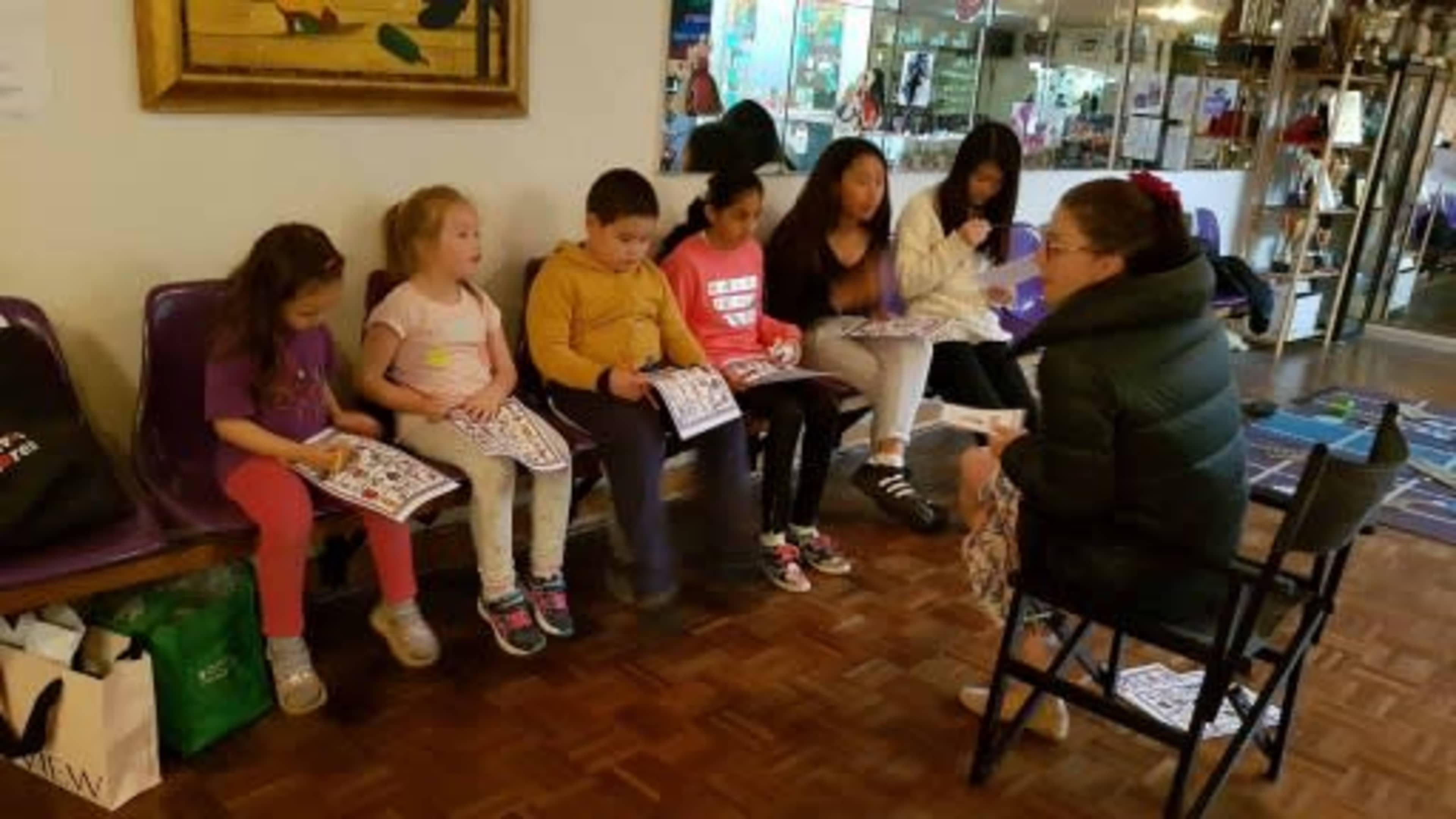 A group of children sit on benches, coloring in books while an adult with their back to the camera speaks to them in a brightly lit room.
