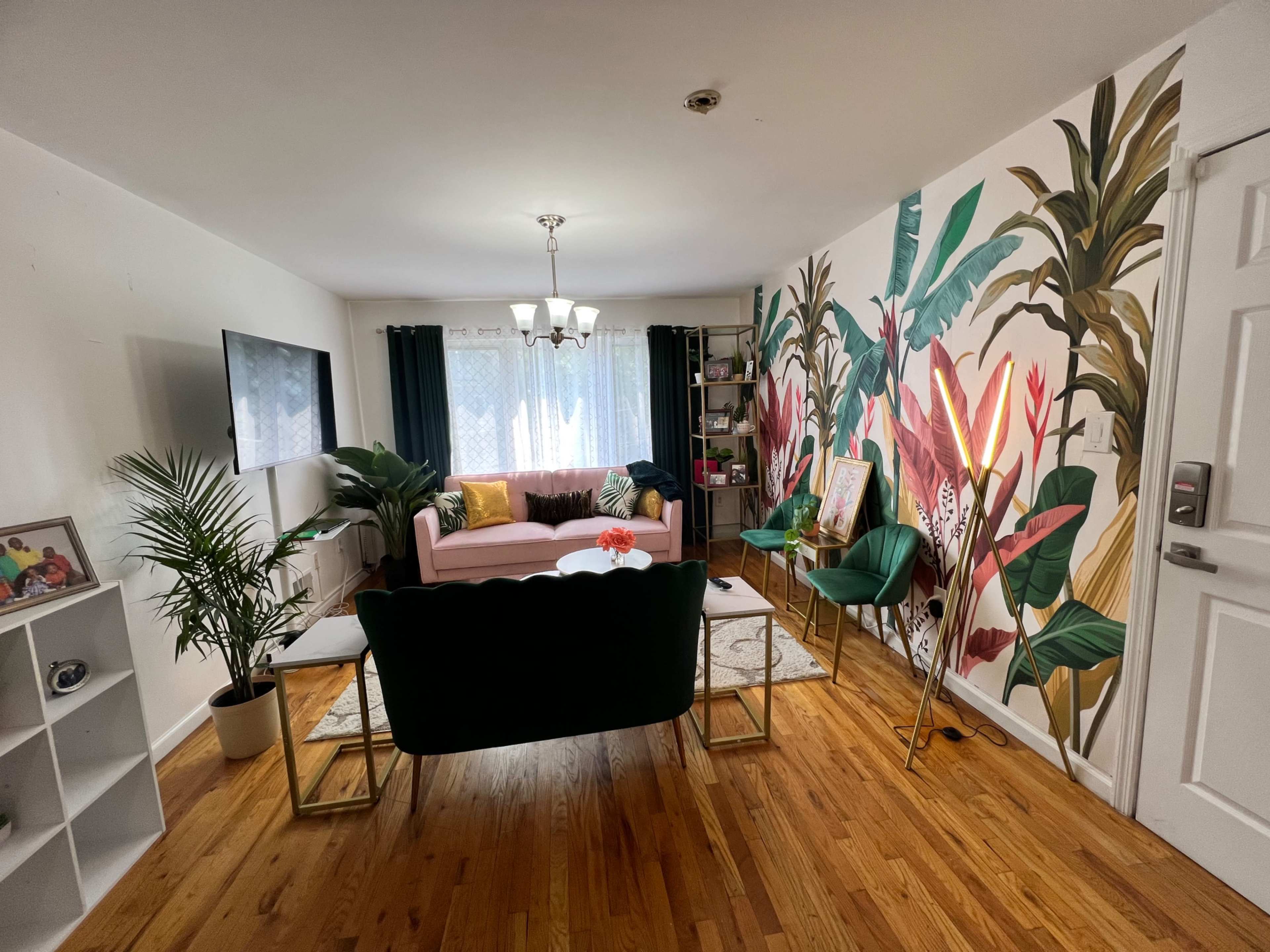 The image shows a living room with a pink sofa, a green armchair, a TV on the wall, and a vibrant plant mural.