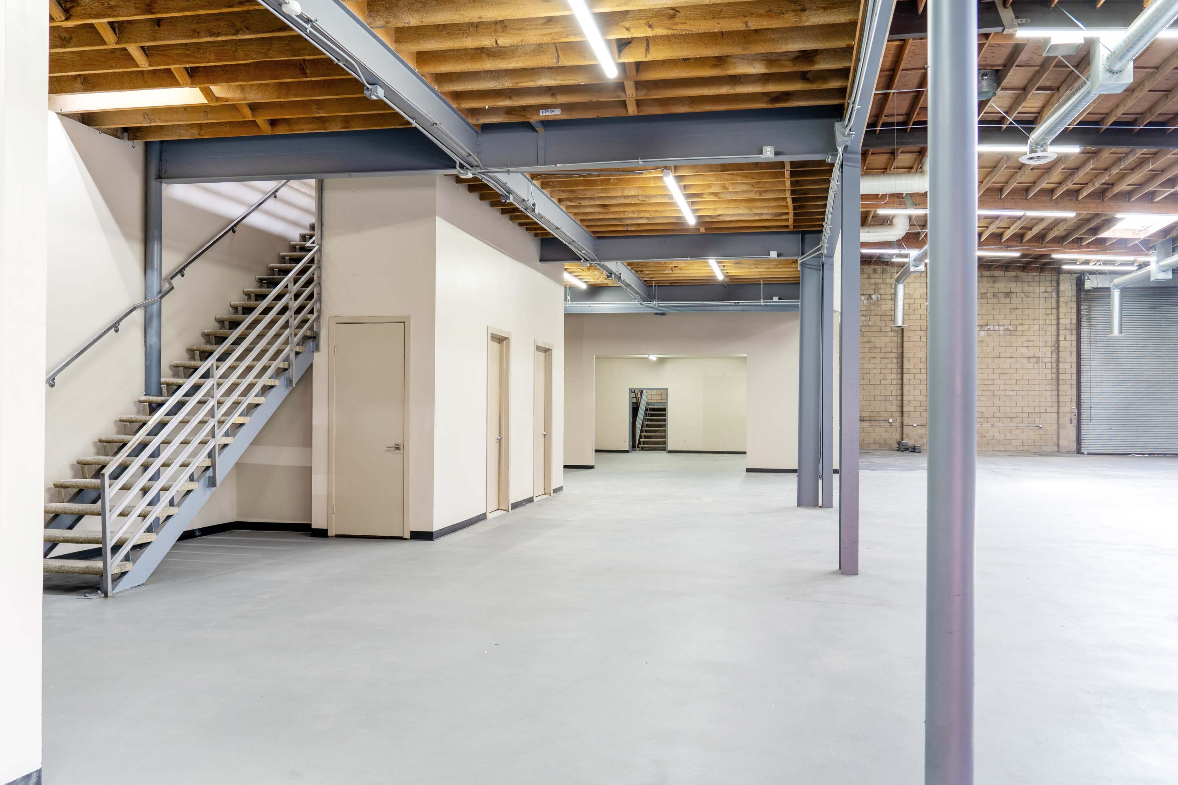 The image shows a spacious, empty interior featuring a staircase, light-colored walls, and exposed wooden beams.