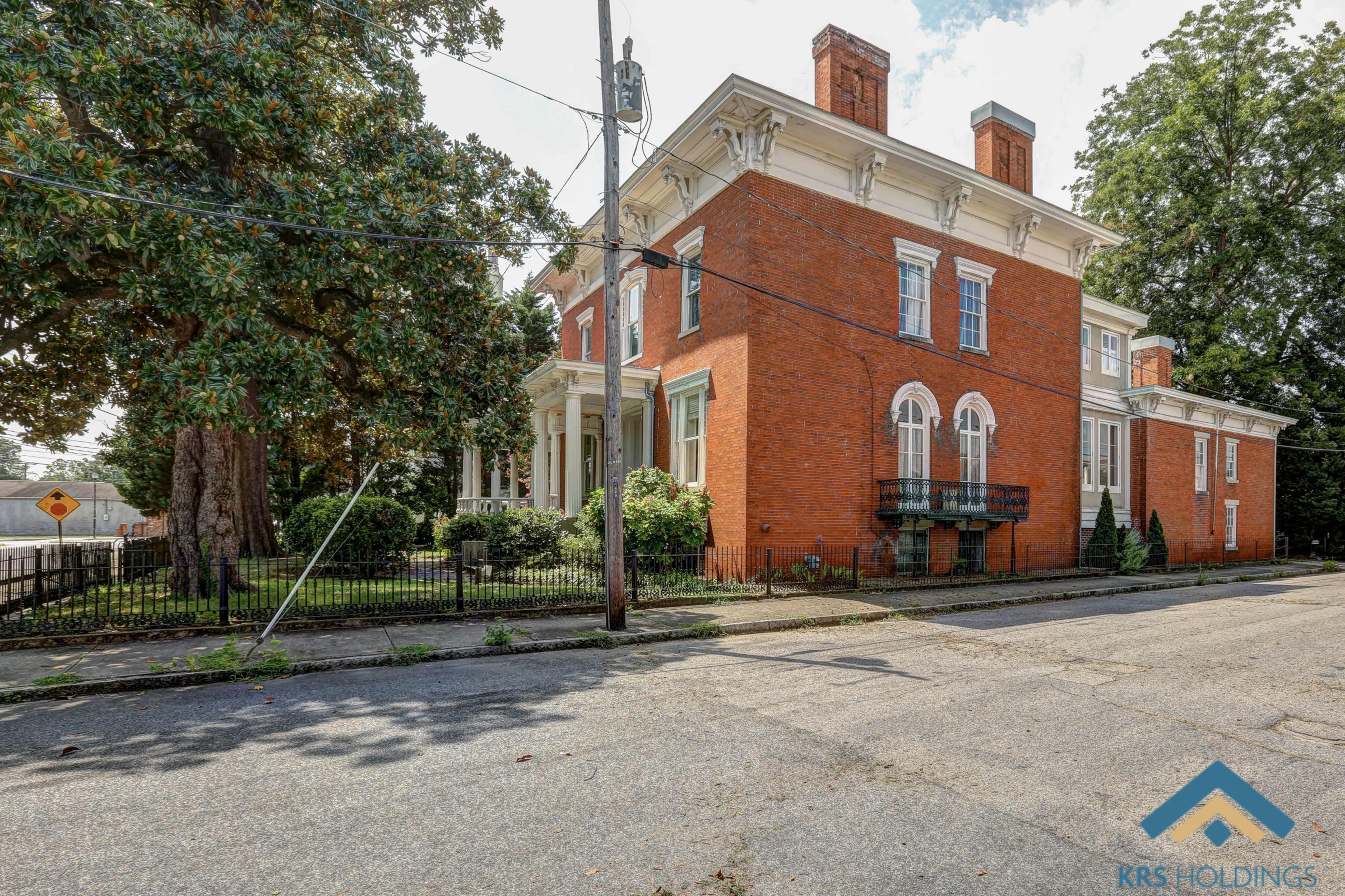 A large, two-story brick house with white trim and a fenced yard, situated on a tree-lined street.