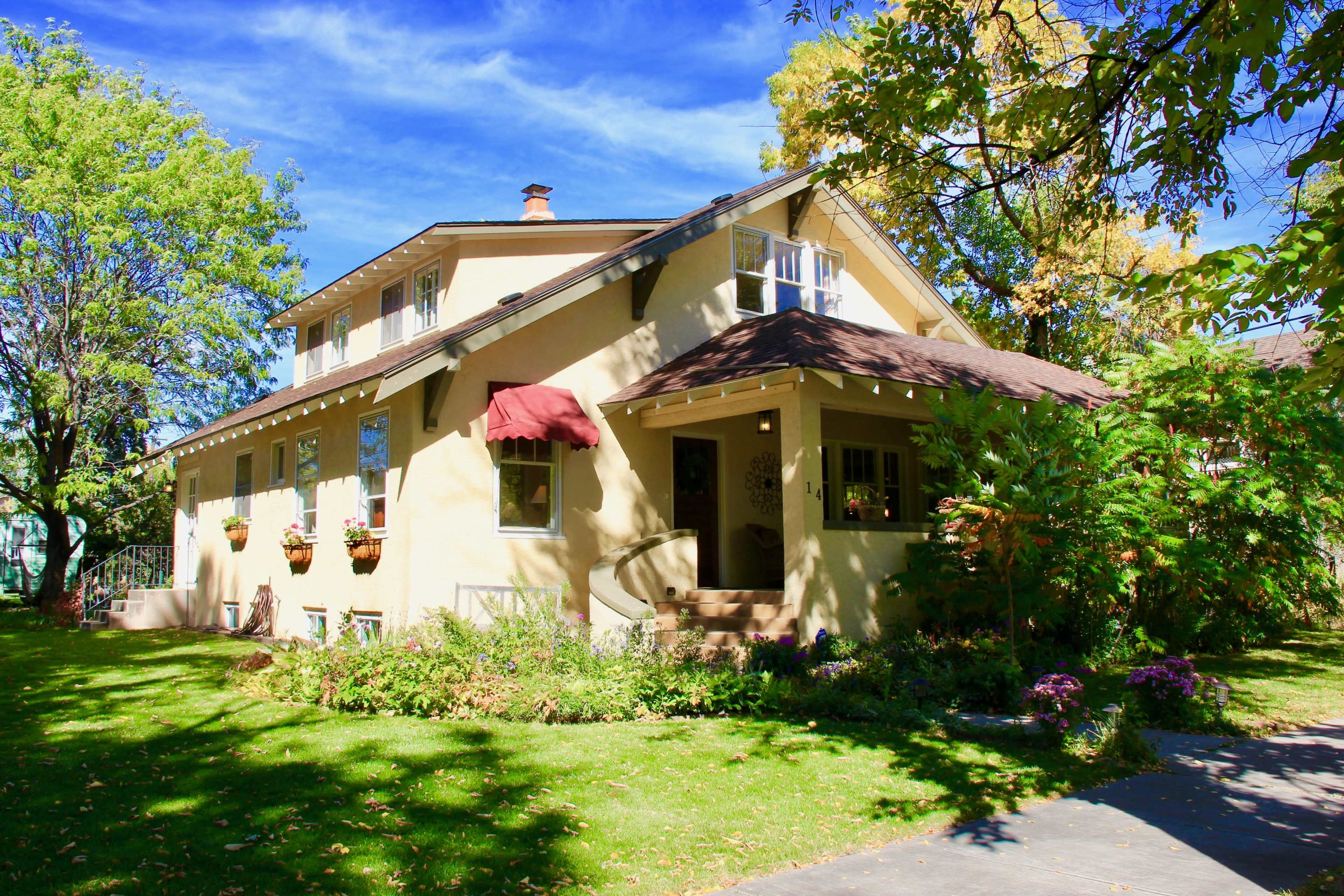 A two-story yellow house with a brown roof and red awning is surrounded by green trees and flower beds.