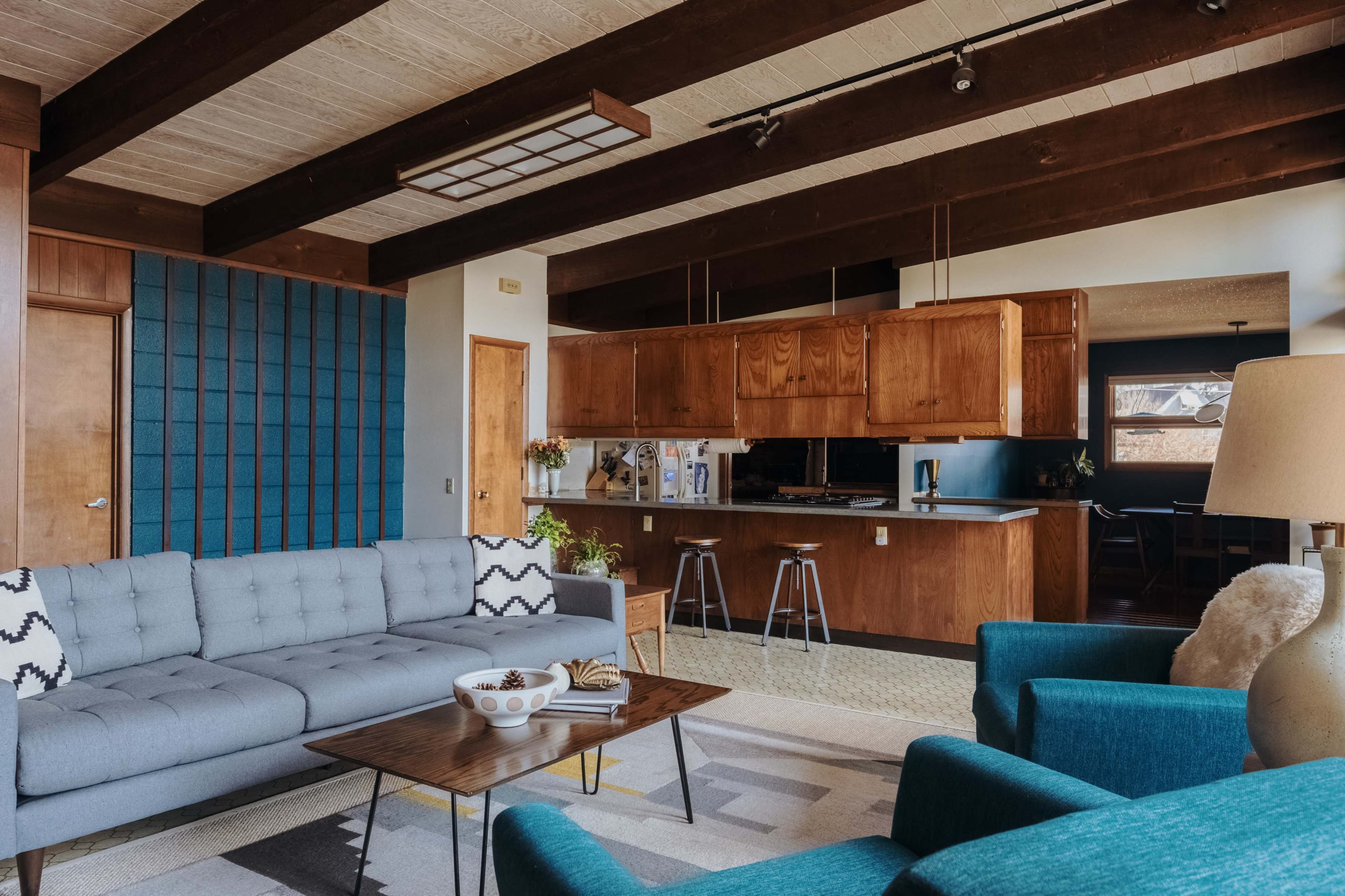 The image shows a modern living room with a blue sofa, teal armchairs, a wooden coffee table, and a kitchen area in the background featuring wood cabinetry and bar stools.