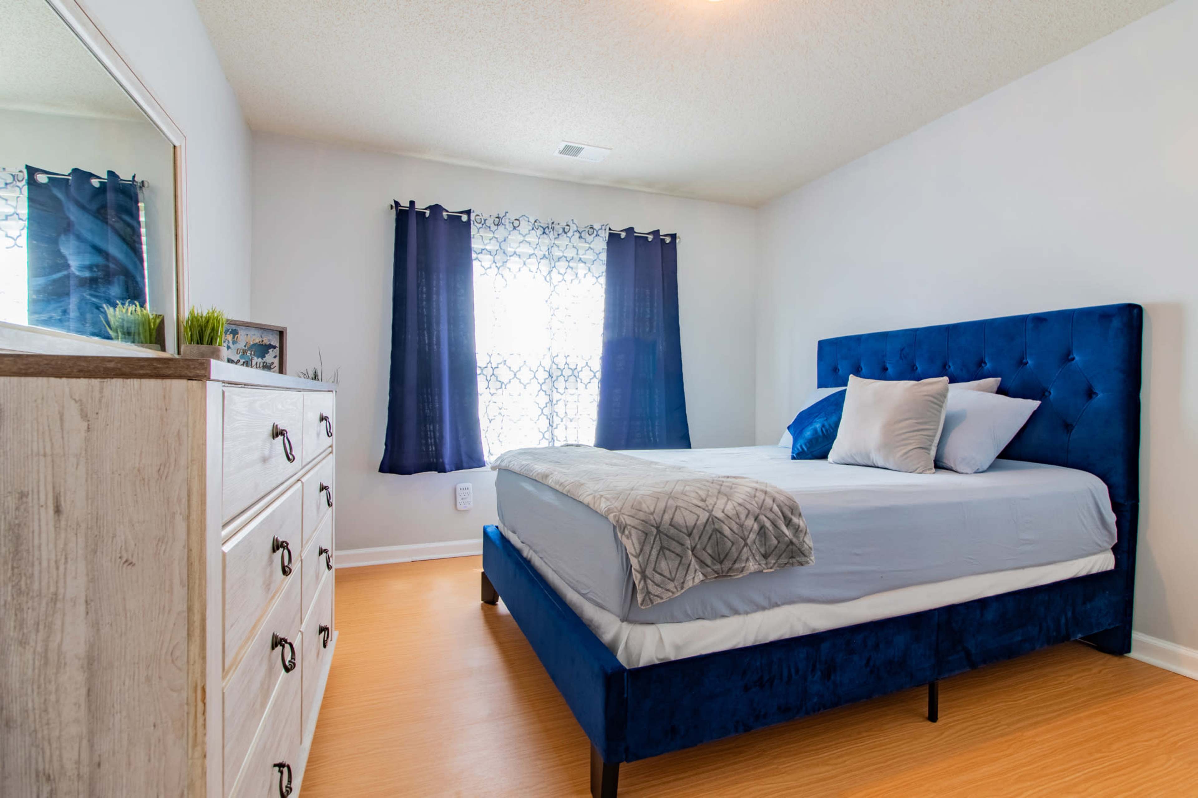 A bedroom with a blue tufted bed, a gray blanket, and a white dresser against a wall, with large windows covered by dark curtains.