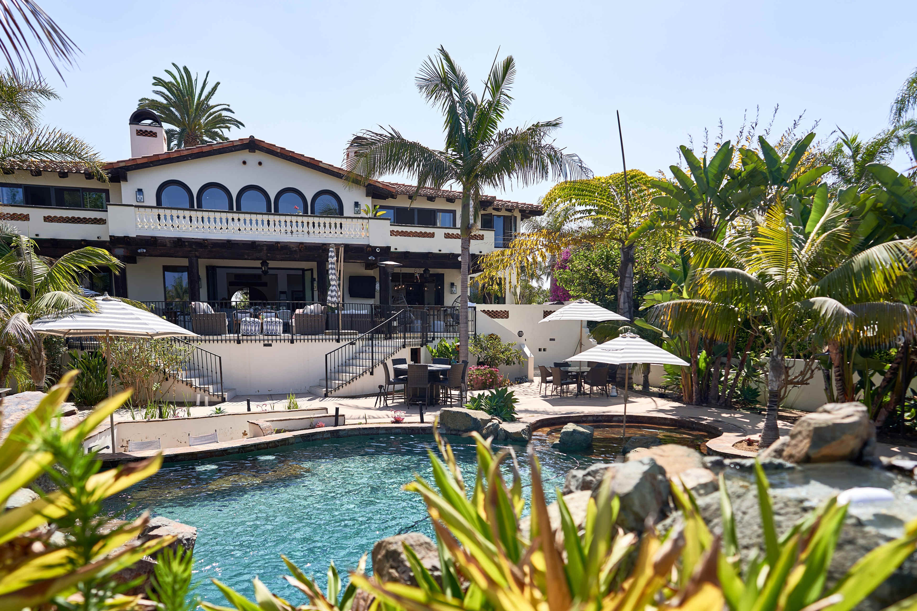 A large house with a balcony and palm trees is situated next to a swimming pool surrounded by rocks and tropical plants.