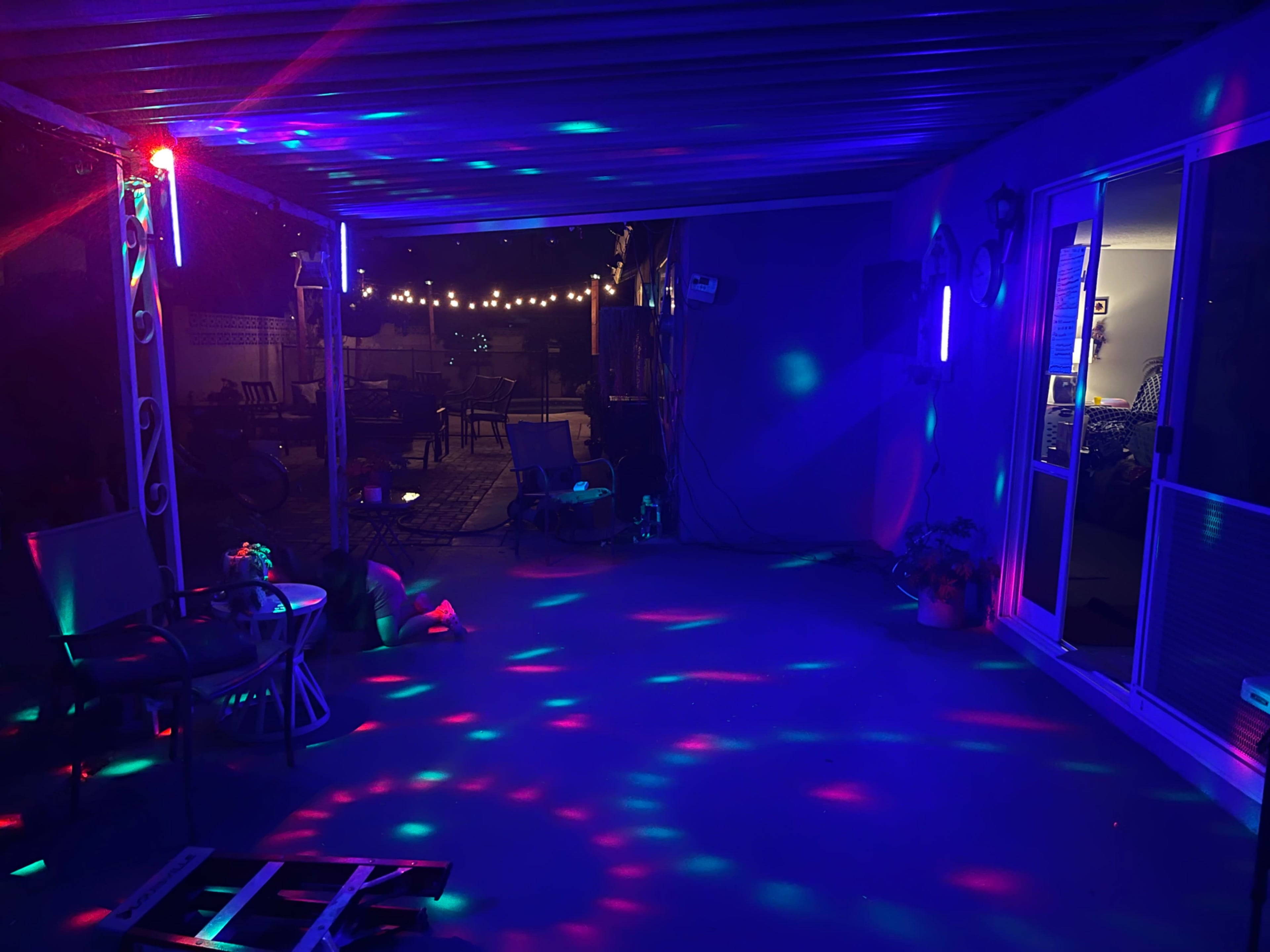 A patio area is illuminated with colorful lights casting patterns on the floor, while string lights hang above in the background.
