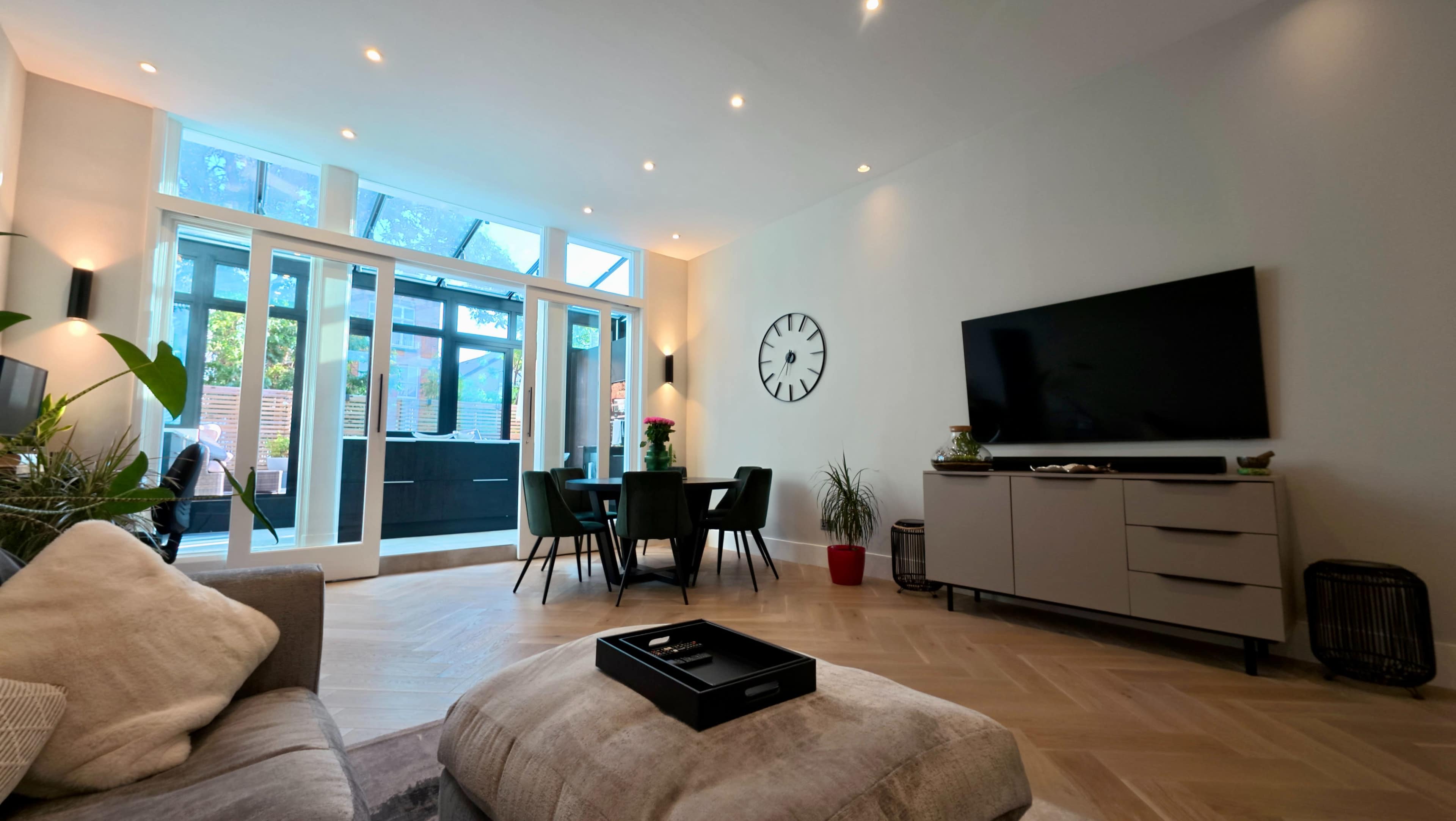 The image shows a modern living room with a large television, a dining area with green chairs, and large glass doors leading to an outdoor space.
