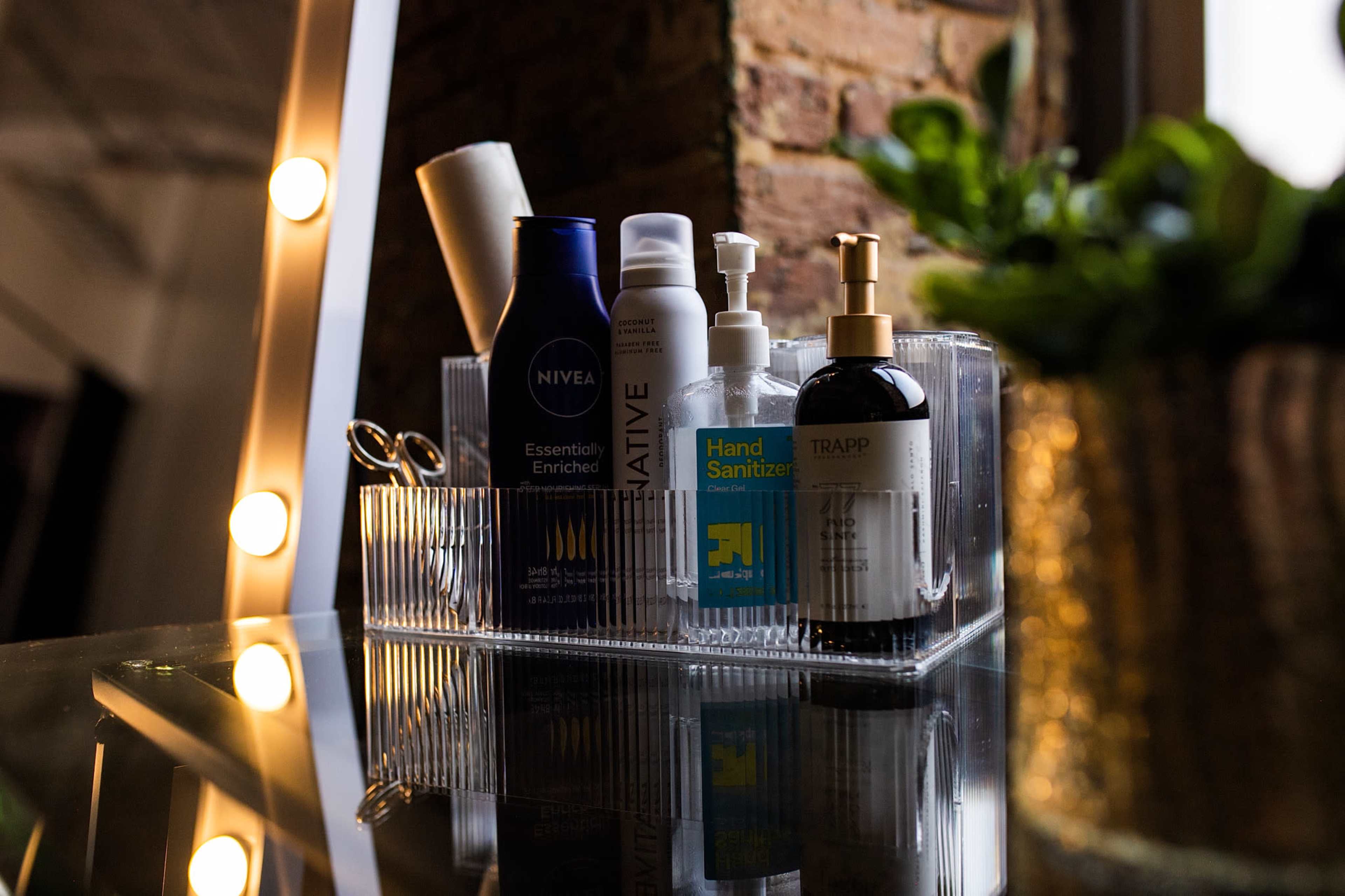 A clear organizer holds various personal care products, including moisturizers and hand sanitizer, on a reflective surface near a window with greenery.