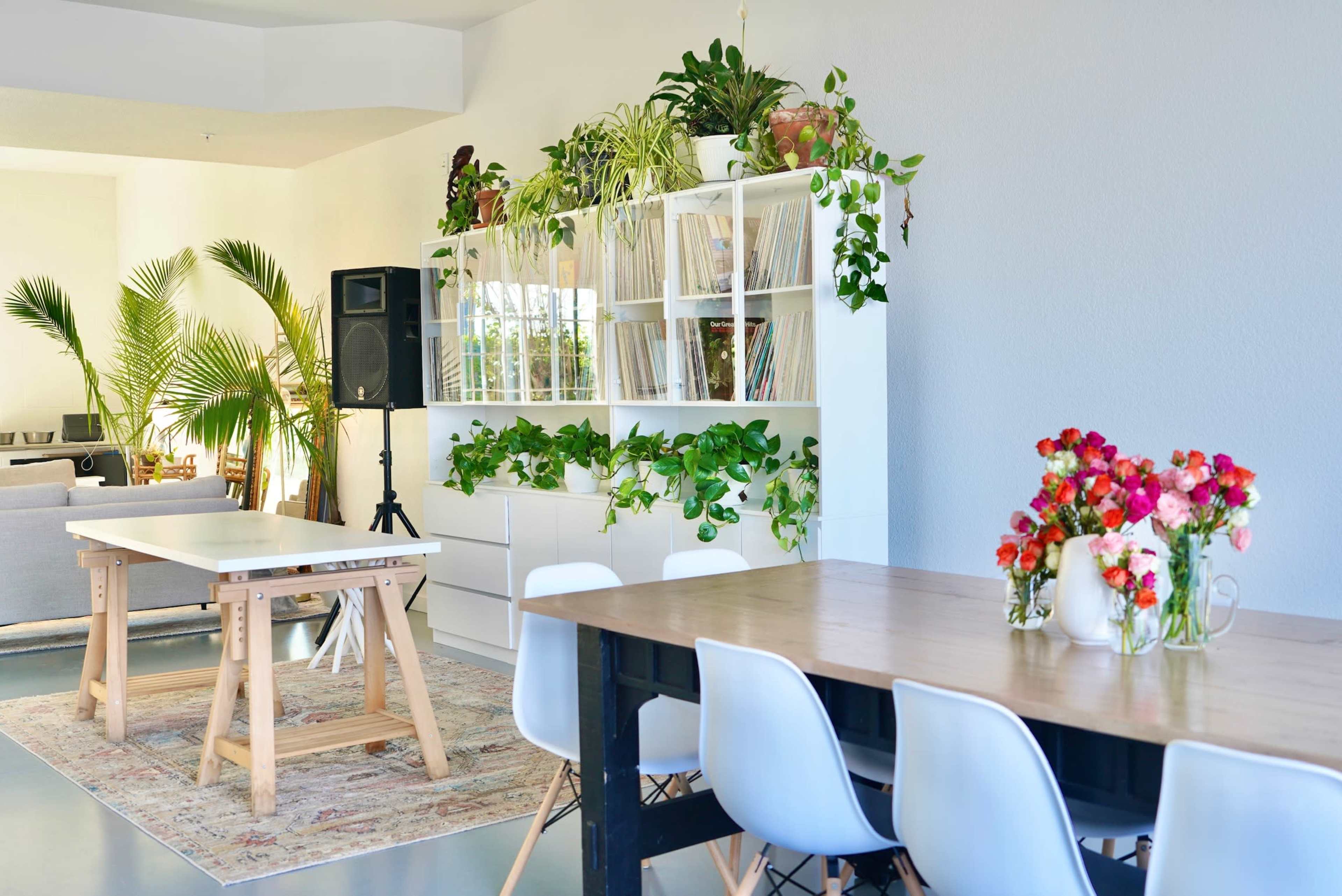 The image shows a bright room with two wooden tables, a shelving unit filled with plants and records, and a speaker in the corner.