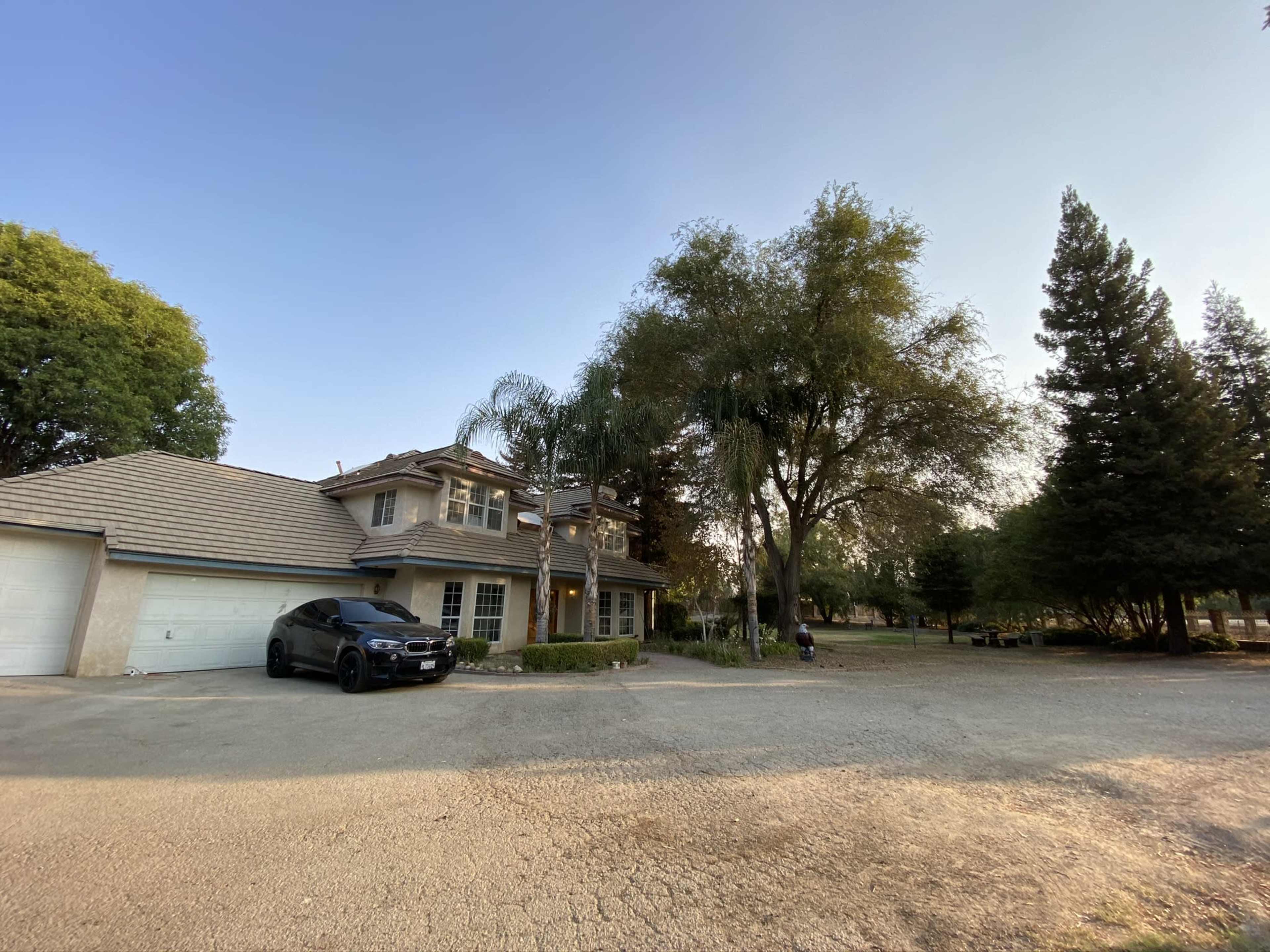 A two-story house with a tiled roof and a black car parked in the driveway is surrounded by trees and a gravel pathway.