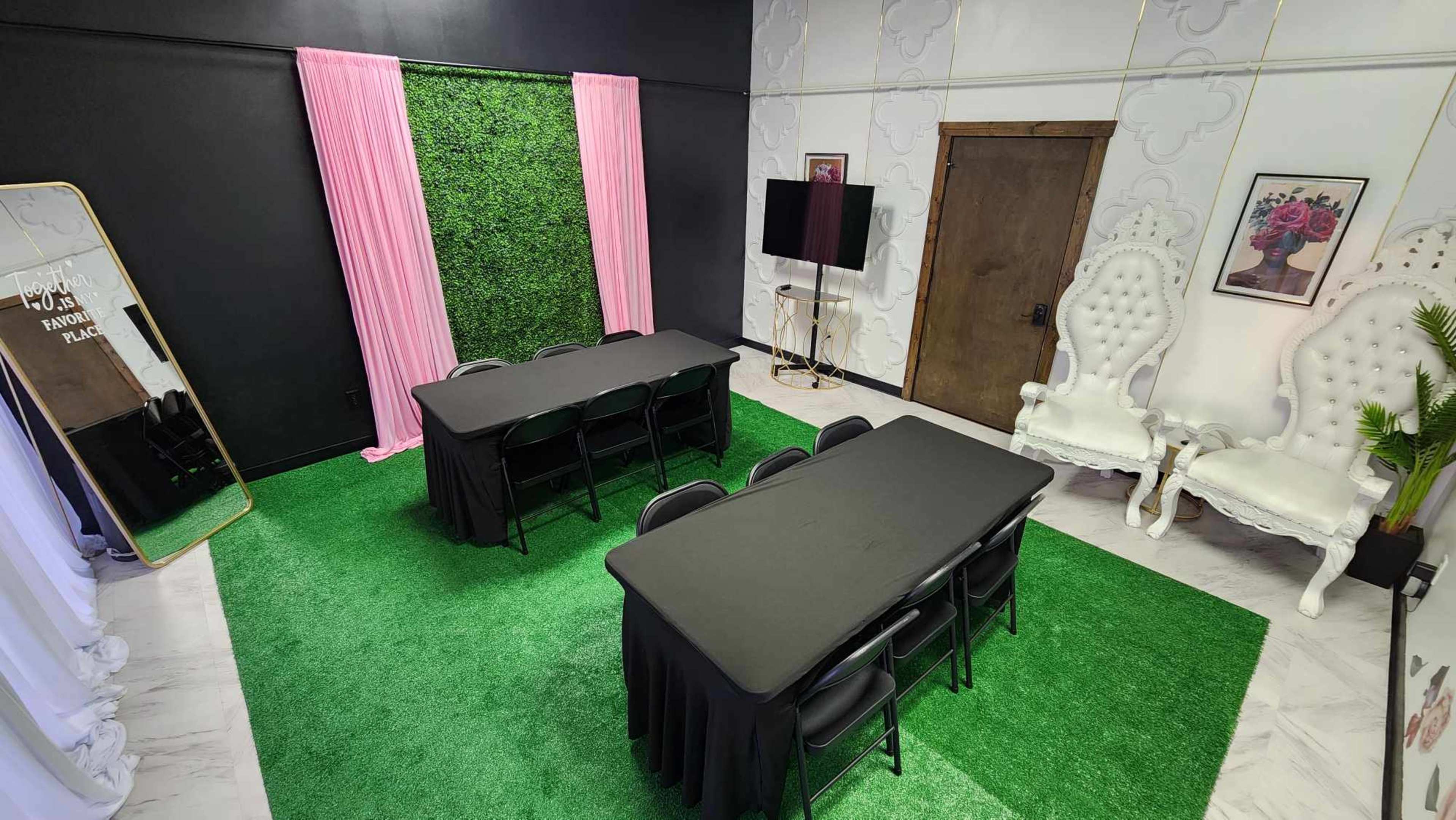 A room with two black tables and chairs on green carpet, flanked by pink curtains and a decorative wall featuring a mirror and art.
