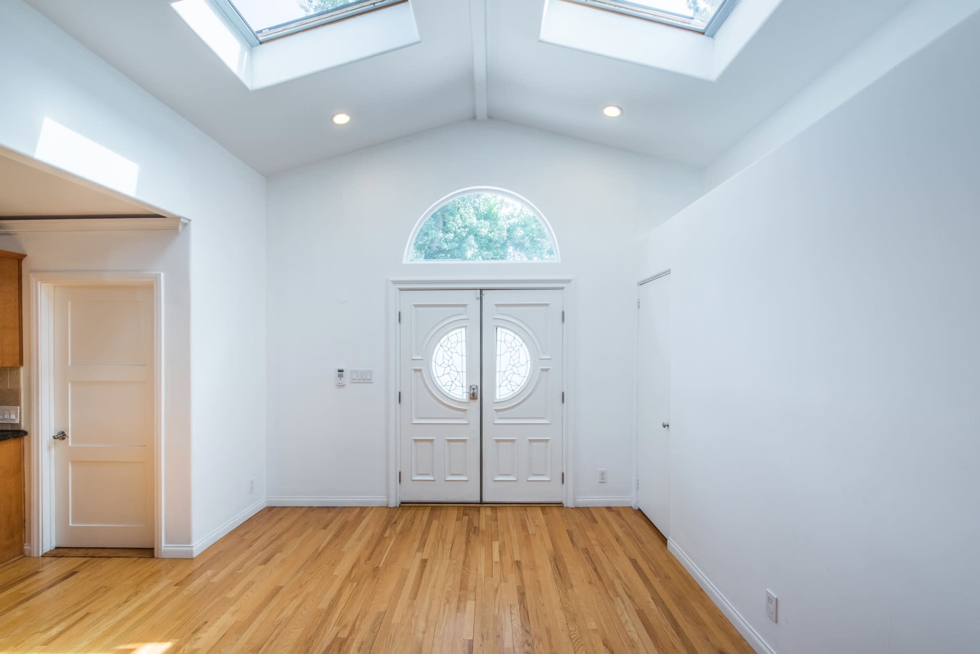 The image shows a bright entryway with a double door featuring circular windows, wooden flooring, and vaulted ceilings with skylights.