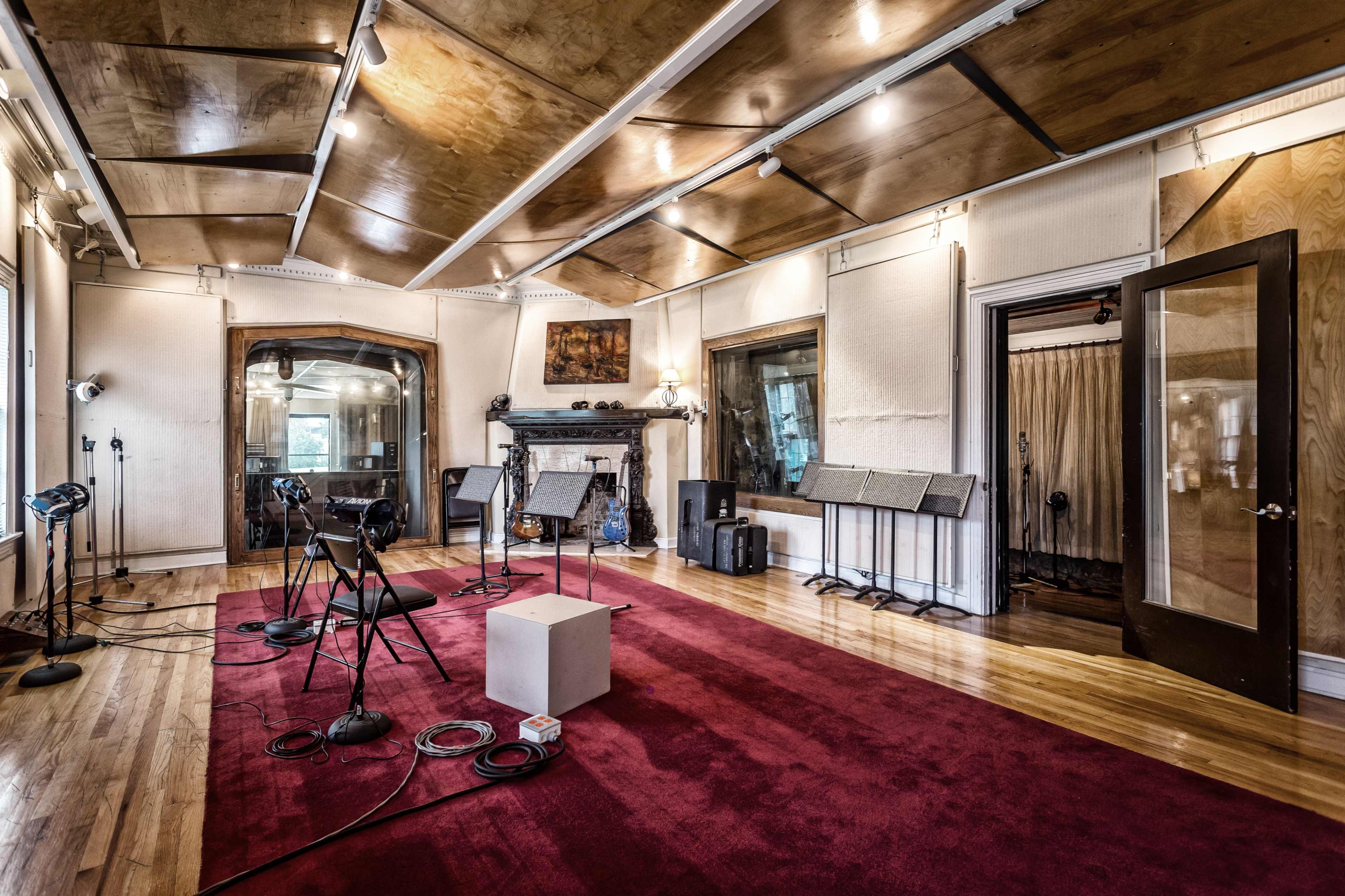 A recording studio features several microphones on stands, music stands, and a small white table on a red carpeted floor with wooden paneling and large windows.