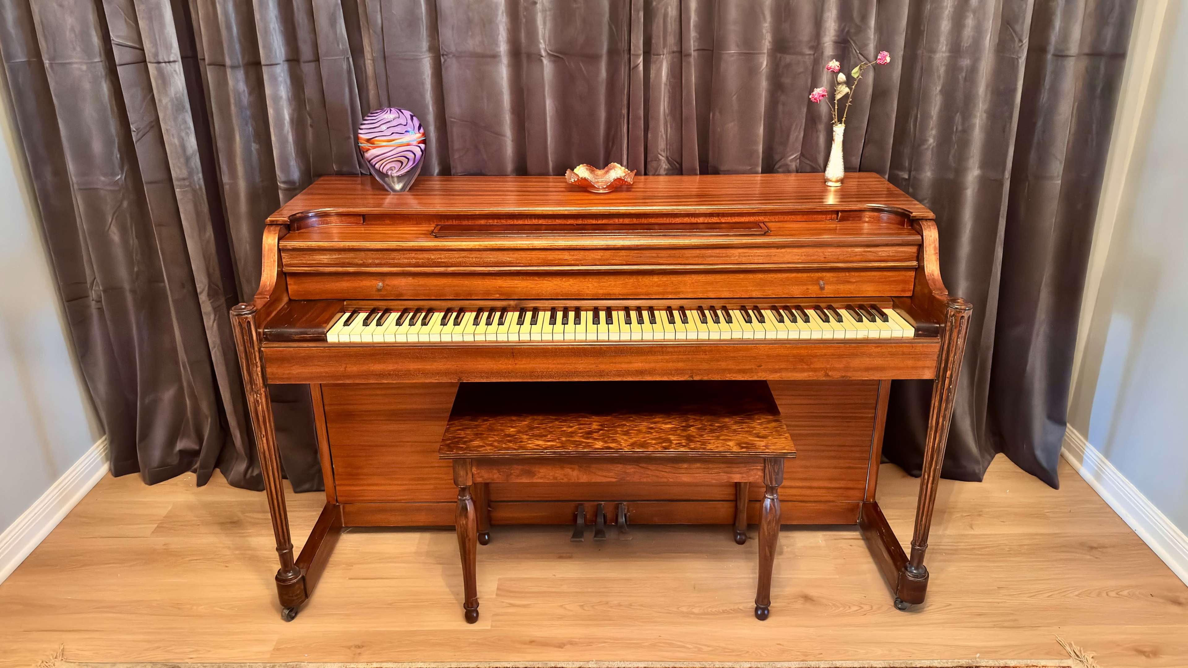 A wooden piano with a matching bench is positioned against a curtain backdrop, complemented by decorative items on top.