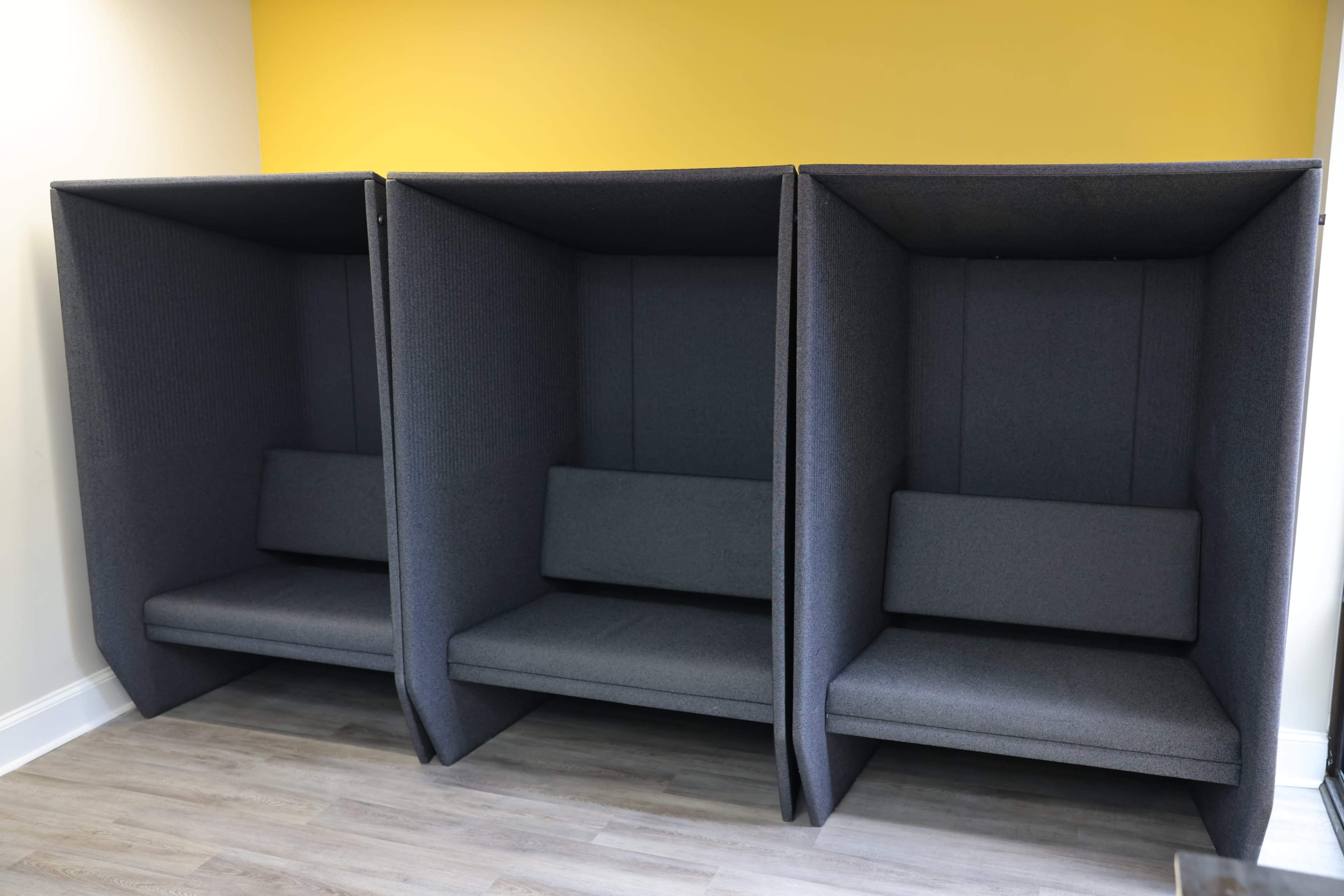 Three individual soundproof booths with grey fabric upholstery and built-in benches, set against a bright yellow wall.
