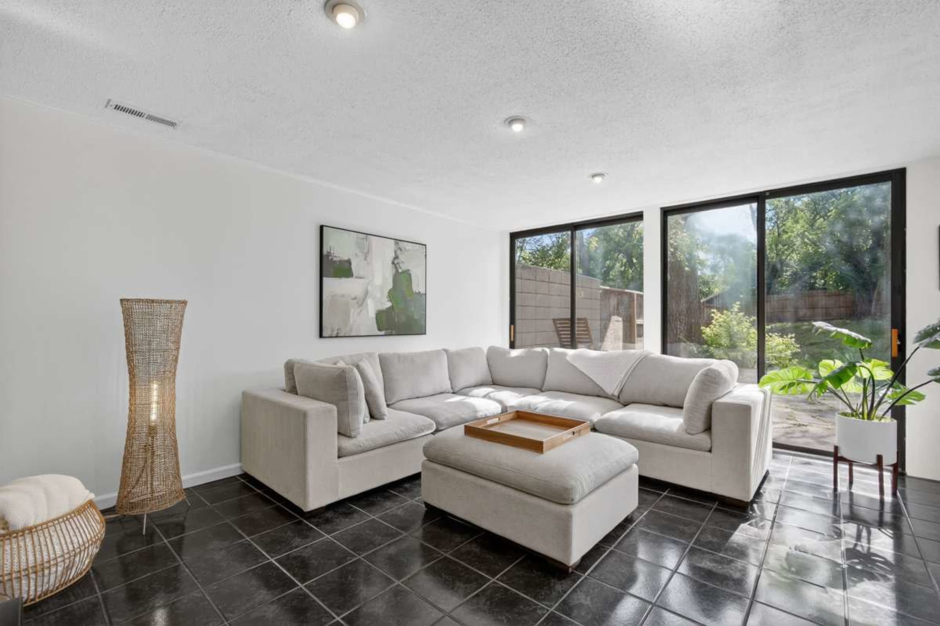 A modern living room features a large sectional sofa, a coffee table, and large windows that provide natural light and a view of a patio and garden.