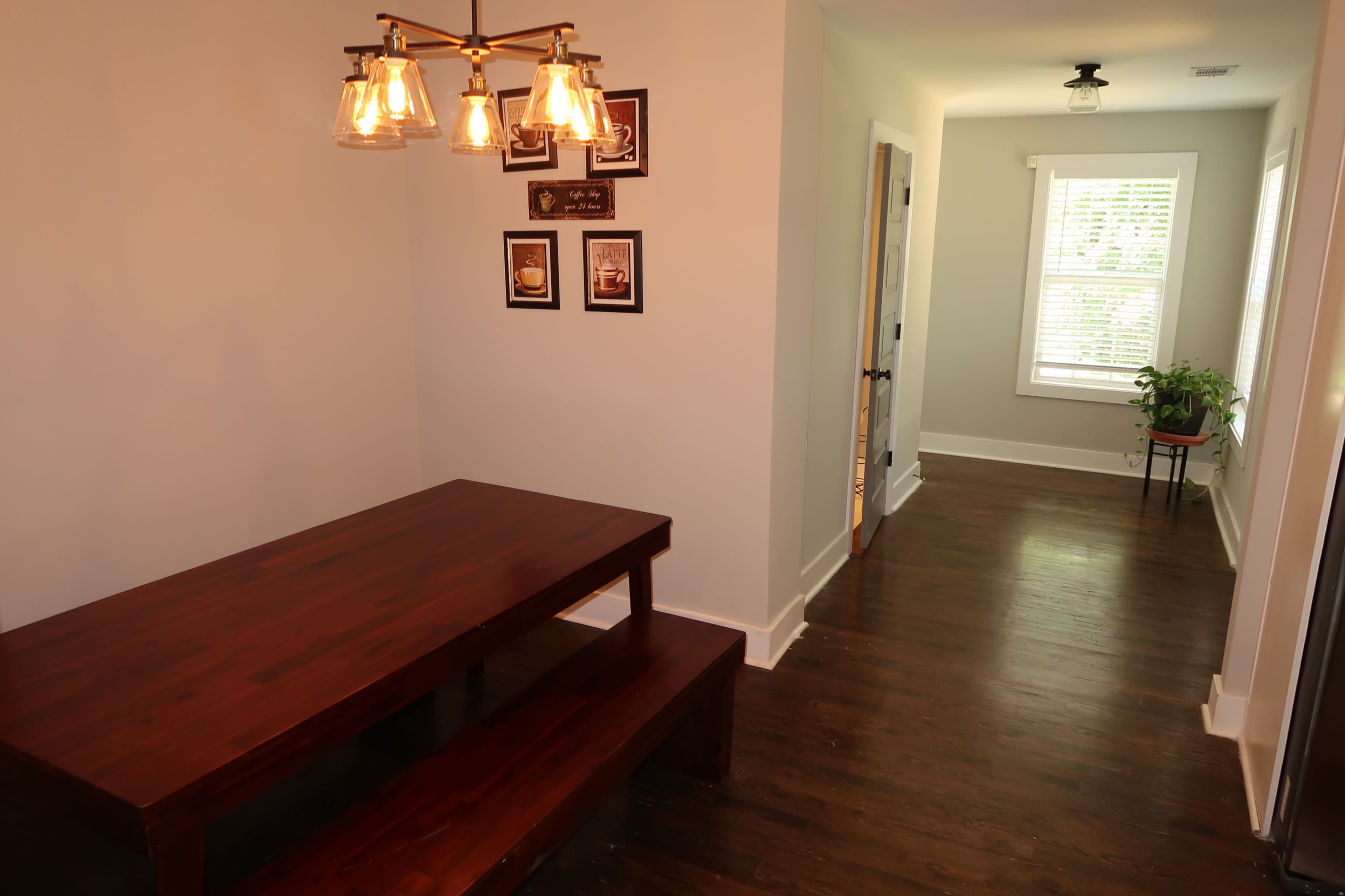 The image depicts a dining area featuring a wooden table with benches and a light fixture above, along with a hallway leading to a room with windows and a potted plant.
