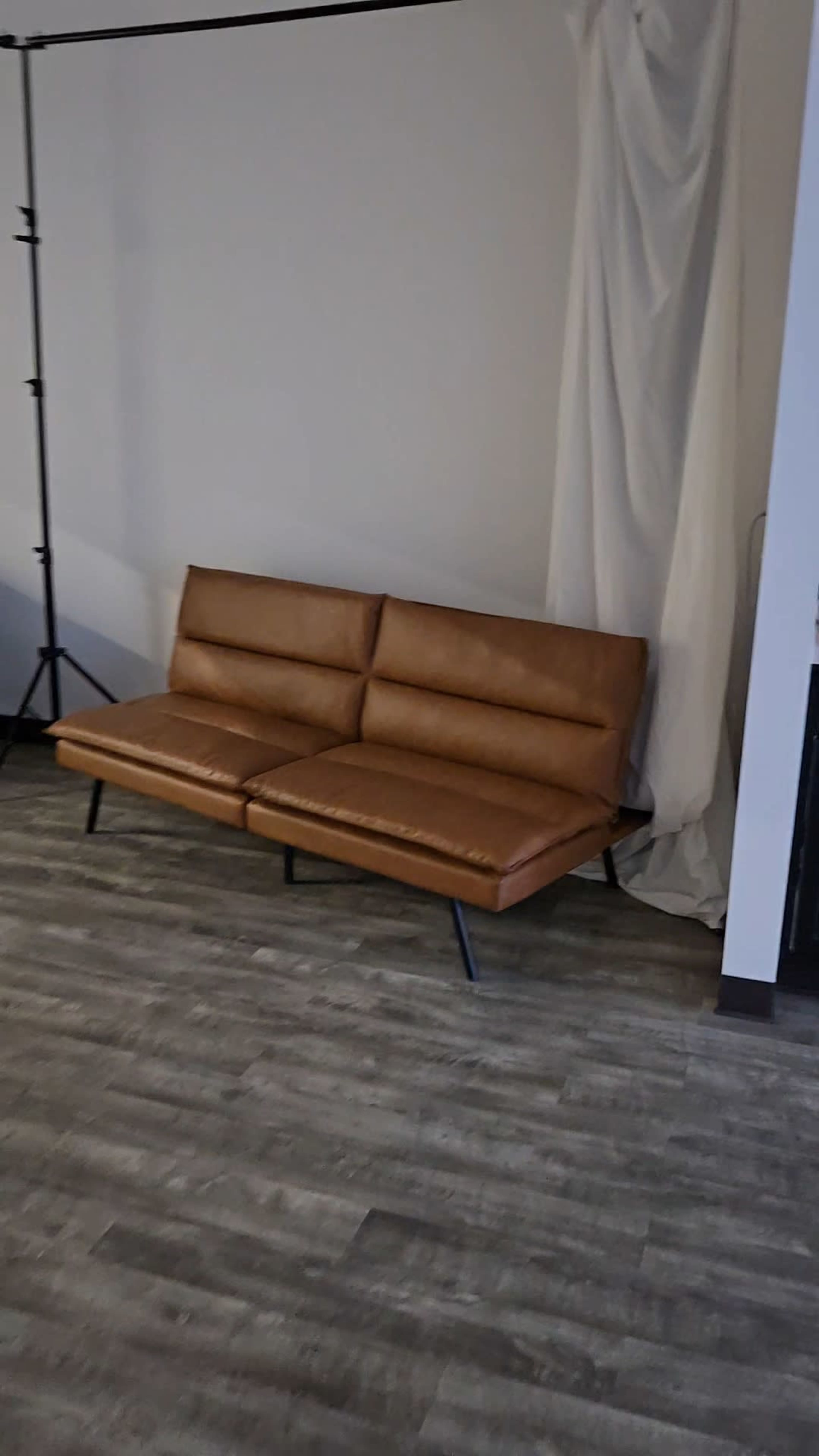 A brown leather couch with sleek metal legs is positioned against a plain wall, next to a draped curtain.