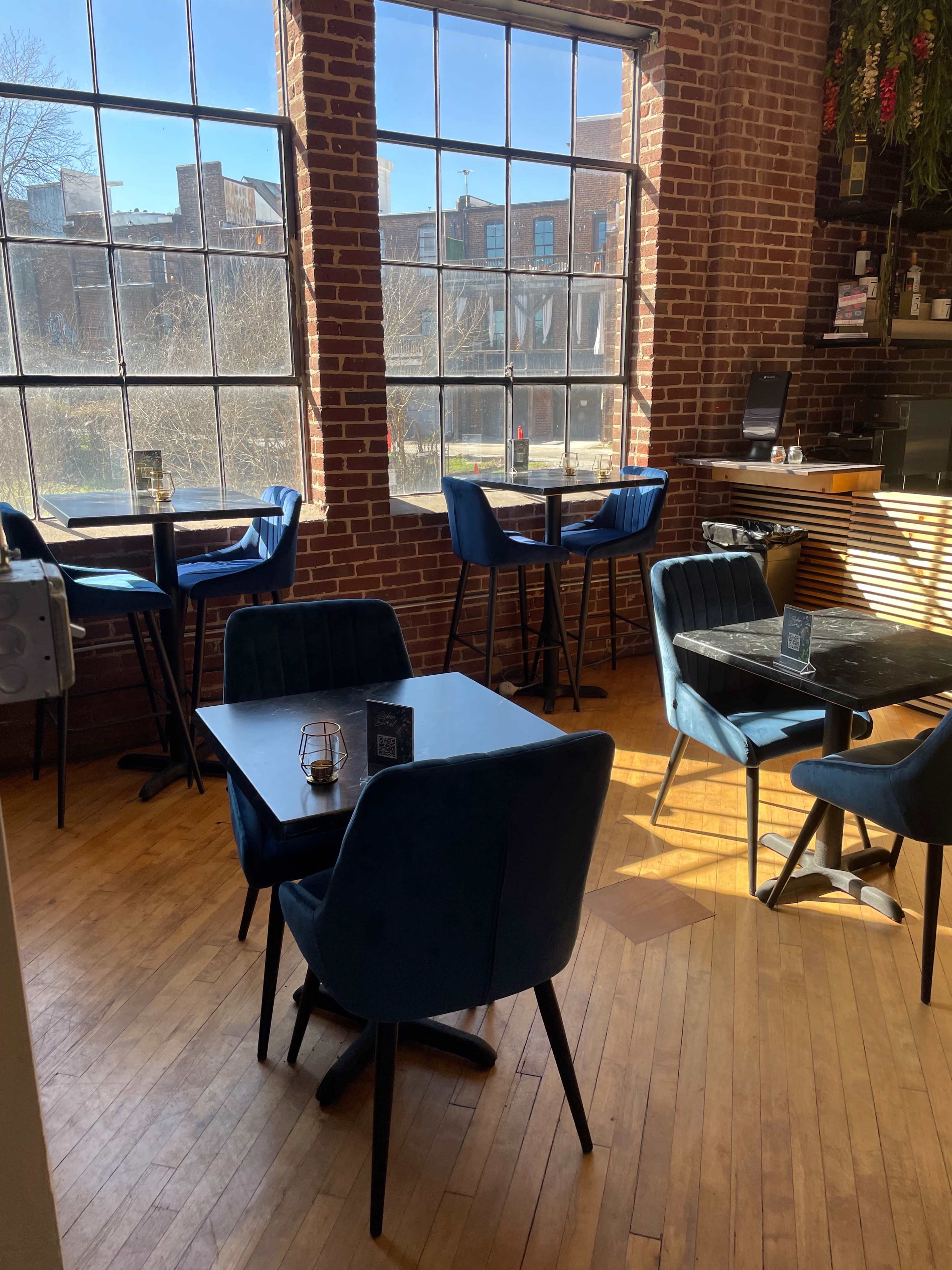 A café interior with brick walls, large windows, and blue upholstered chairs around black tables.