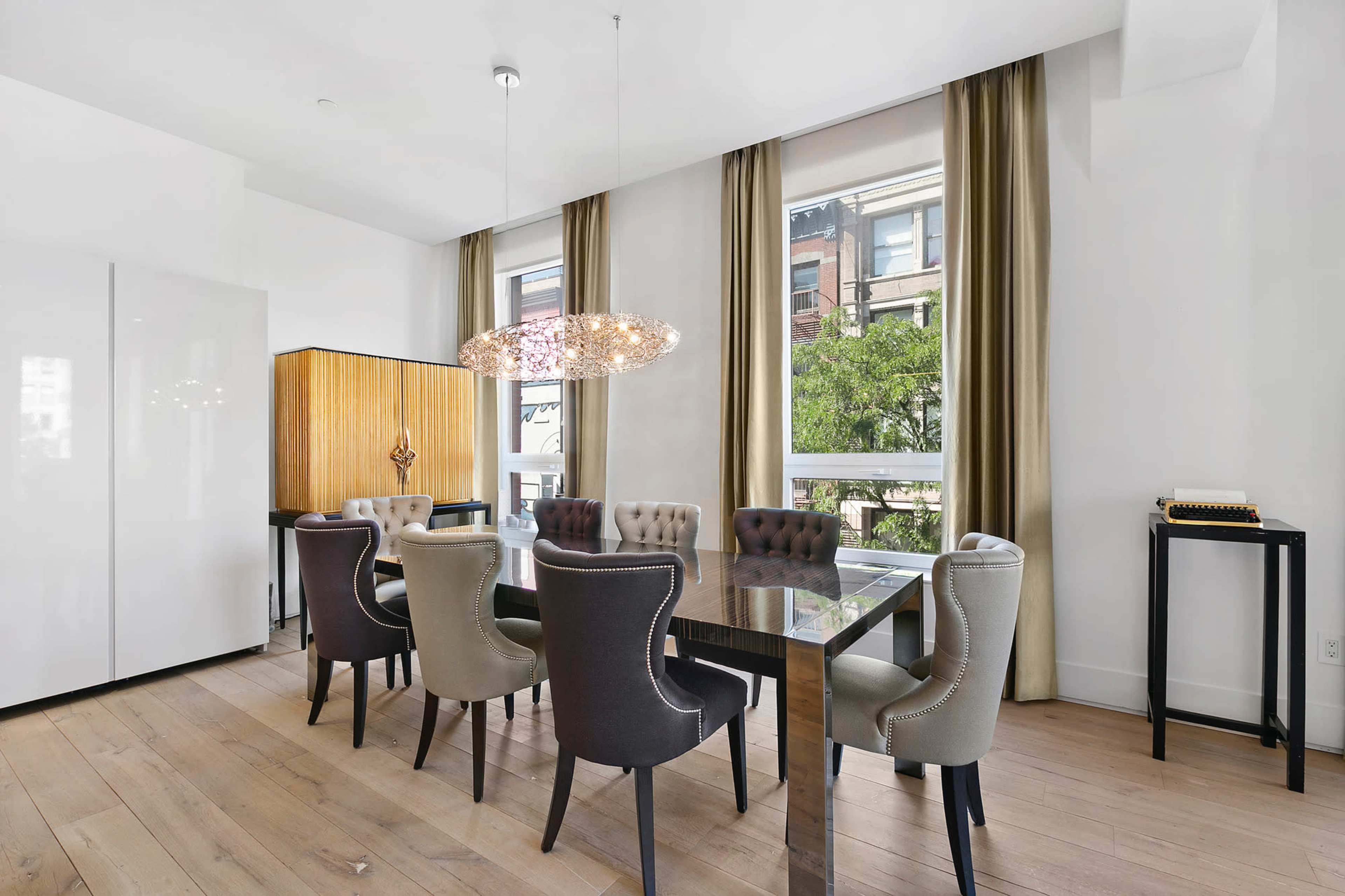 A modern dining area features a rectangular table surrounded by eight upholstered chairs, with large windows allowing natural light to enter the space.