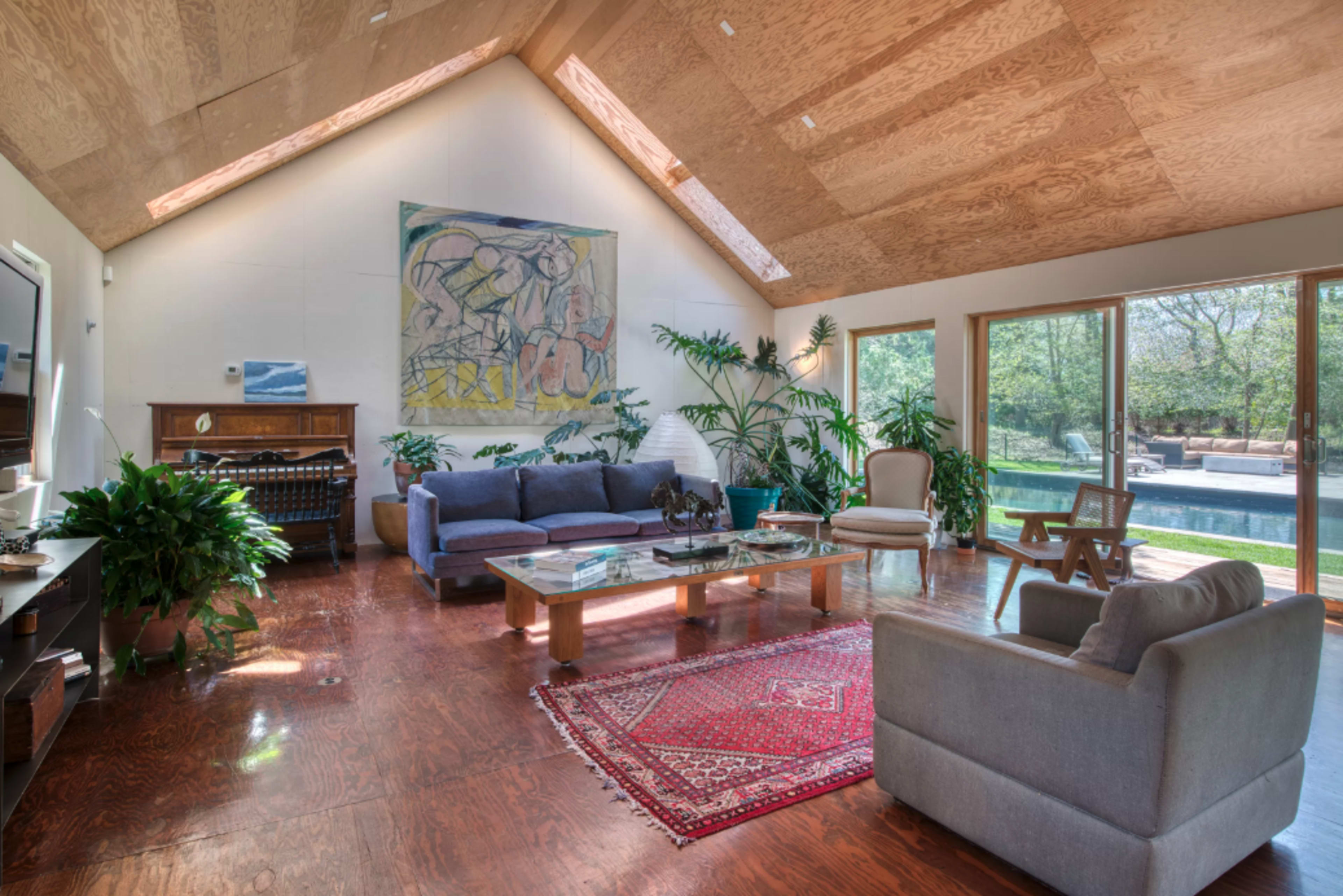 A modern living room with high ceilings, large windows, and a mix of seating arrangements, featuring plants and artwork on the walls.