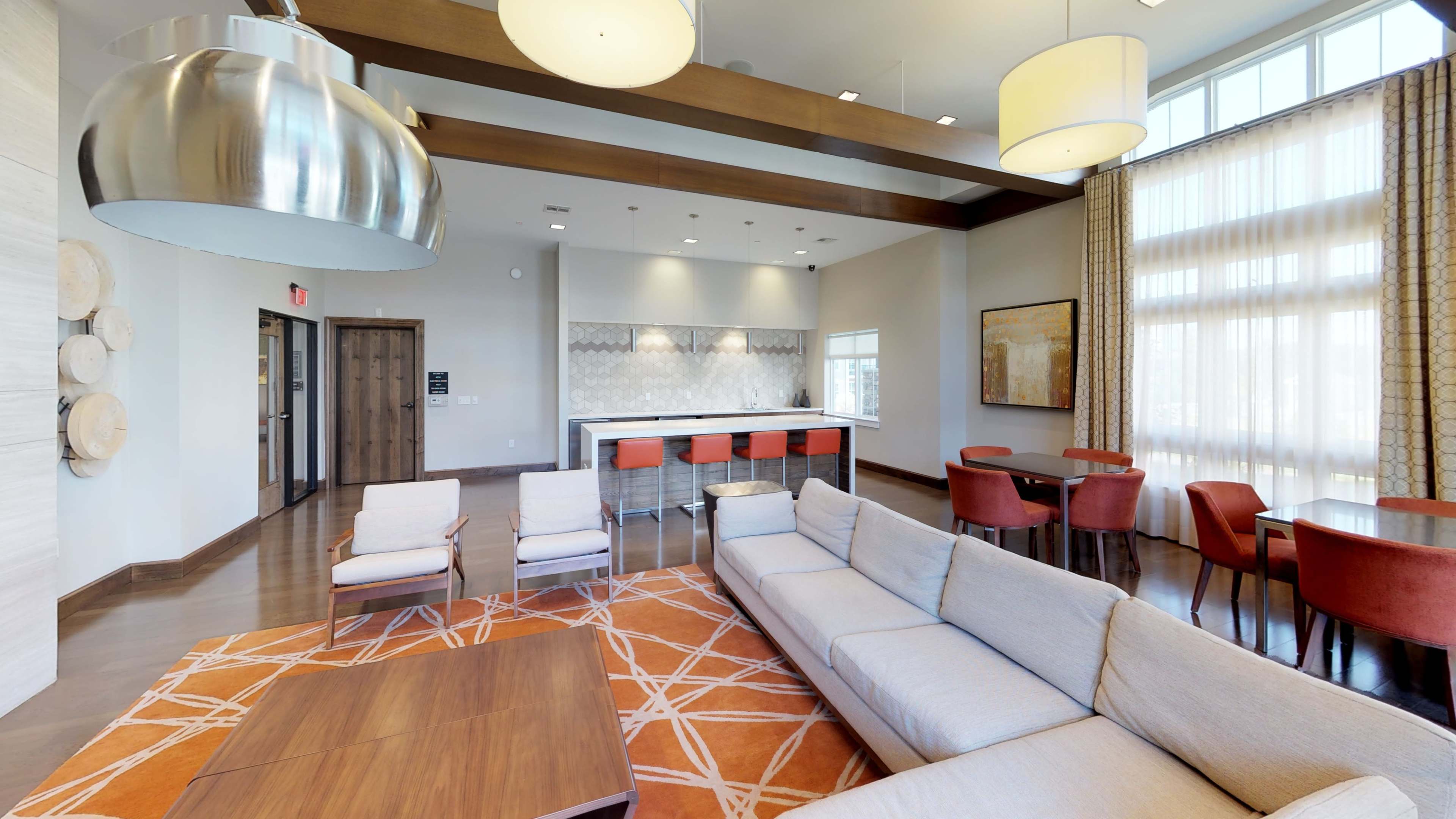 A modern lounge area with a sectional sofa, a coffee table, and a bar with stools, illuminated by pendant lighting and large windows.