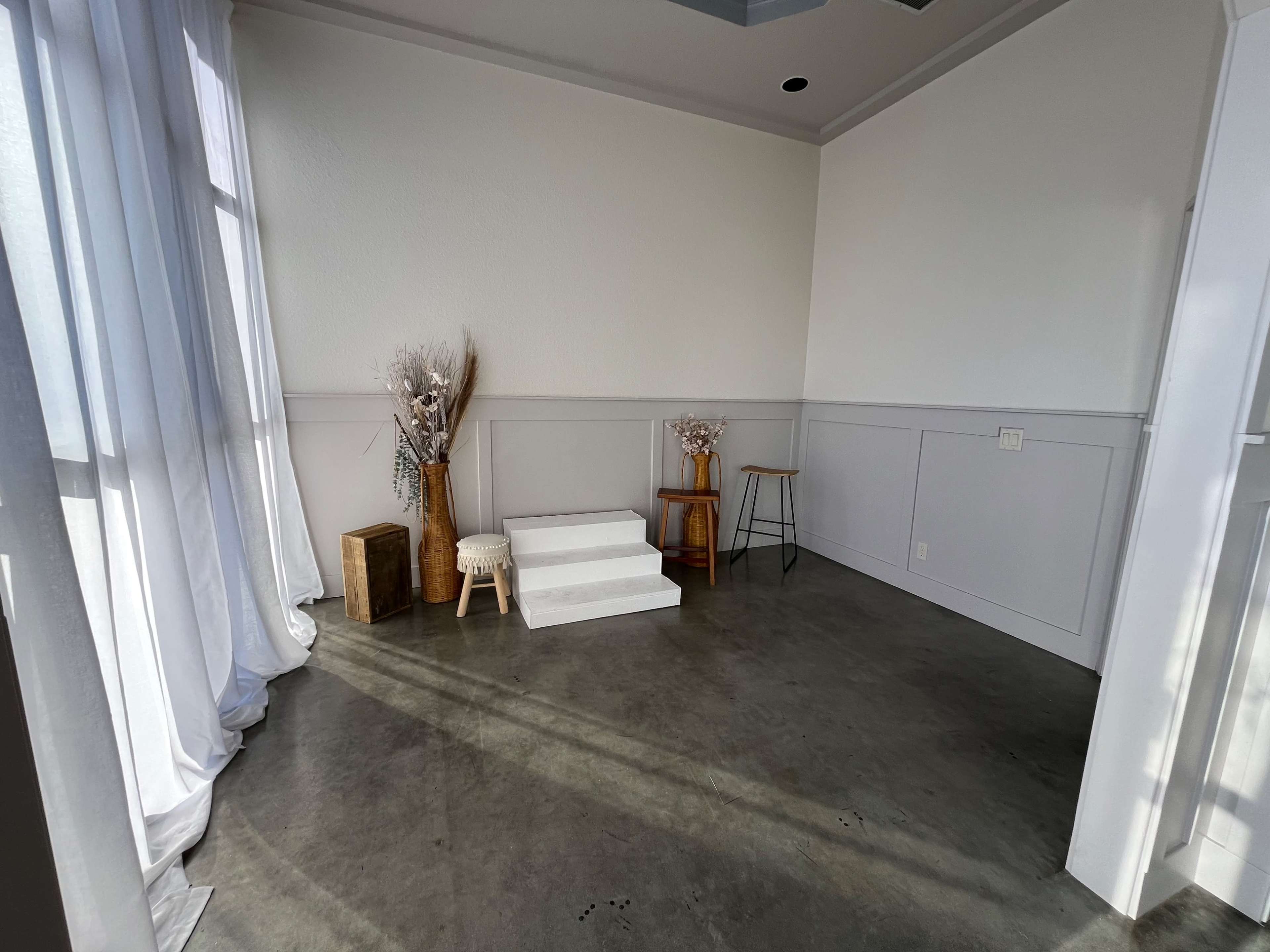 The image shows a minimalist indoor space featuring a small set of white steps, two decorative arrangements, and a stool against a plain wall with large windows allowing natural light to enter.