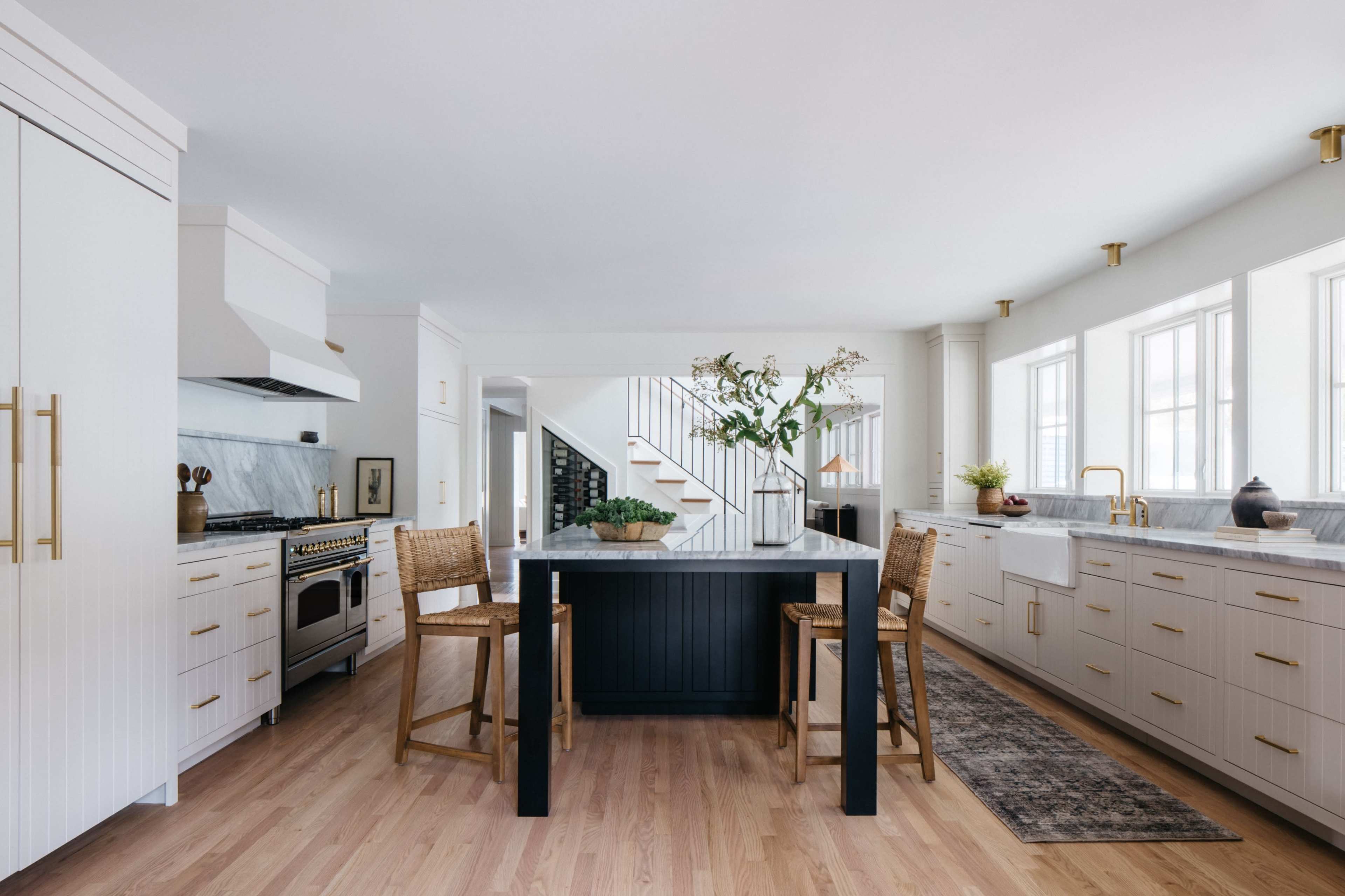 A modern kitchen features a central island with bar stools, white cabinetry, a marble countertop, and large windows allowing natural light.
