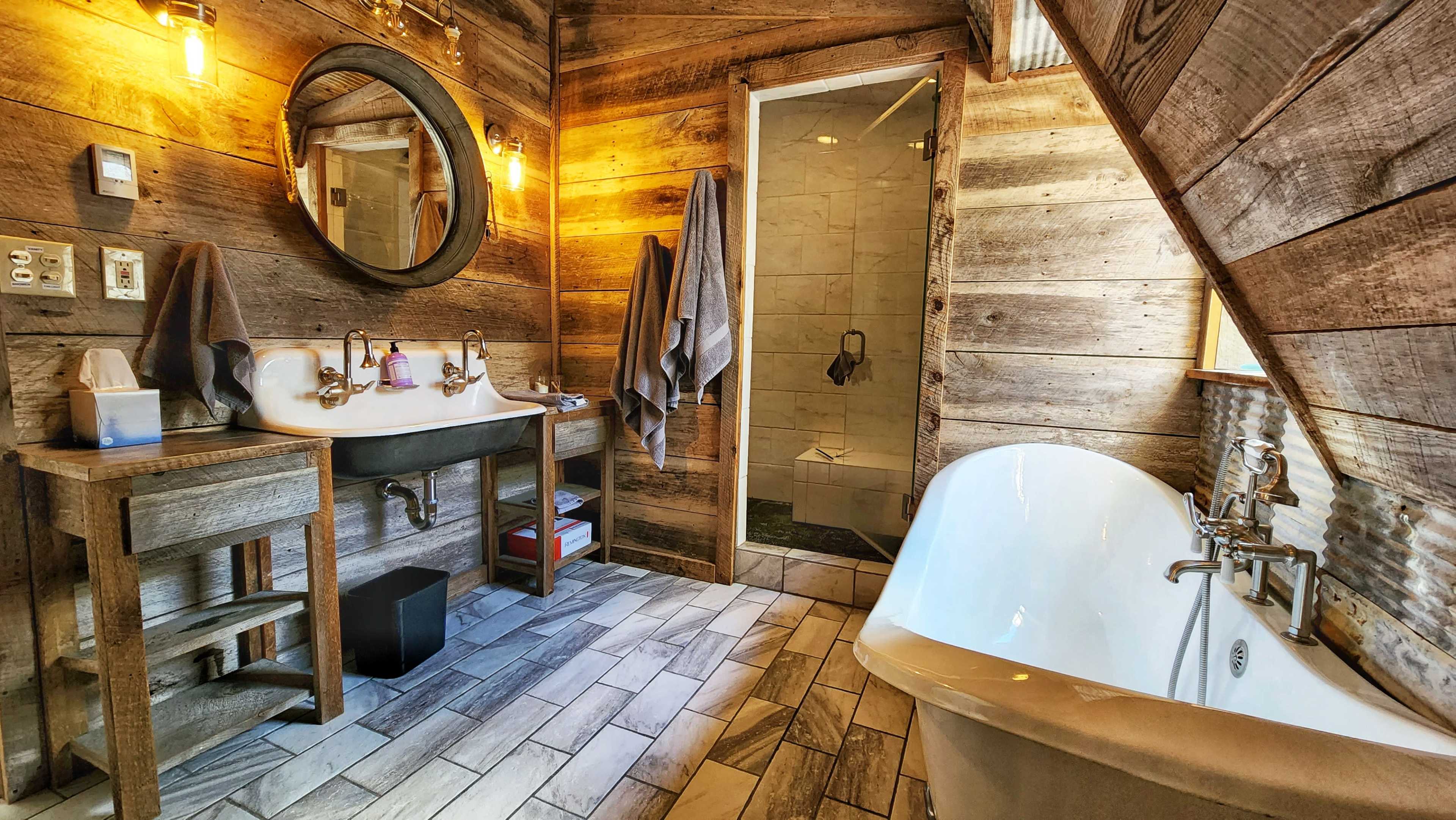 A bathroom featuring a freestanding bathtub, a double sink with a wooden vanity, a glass shower, and rustic wooden walls.