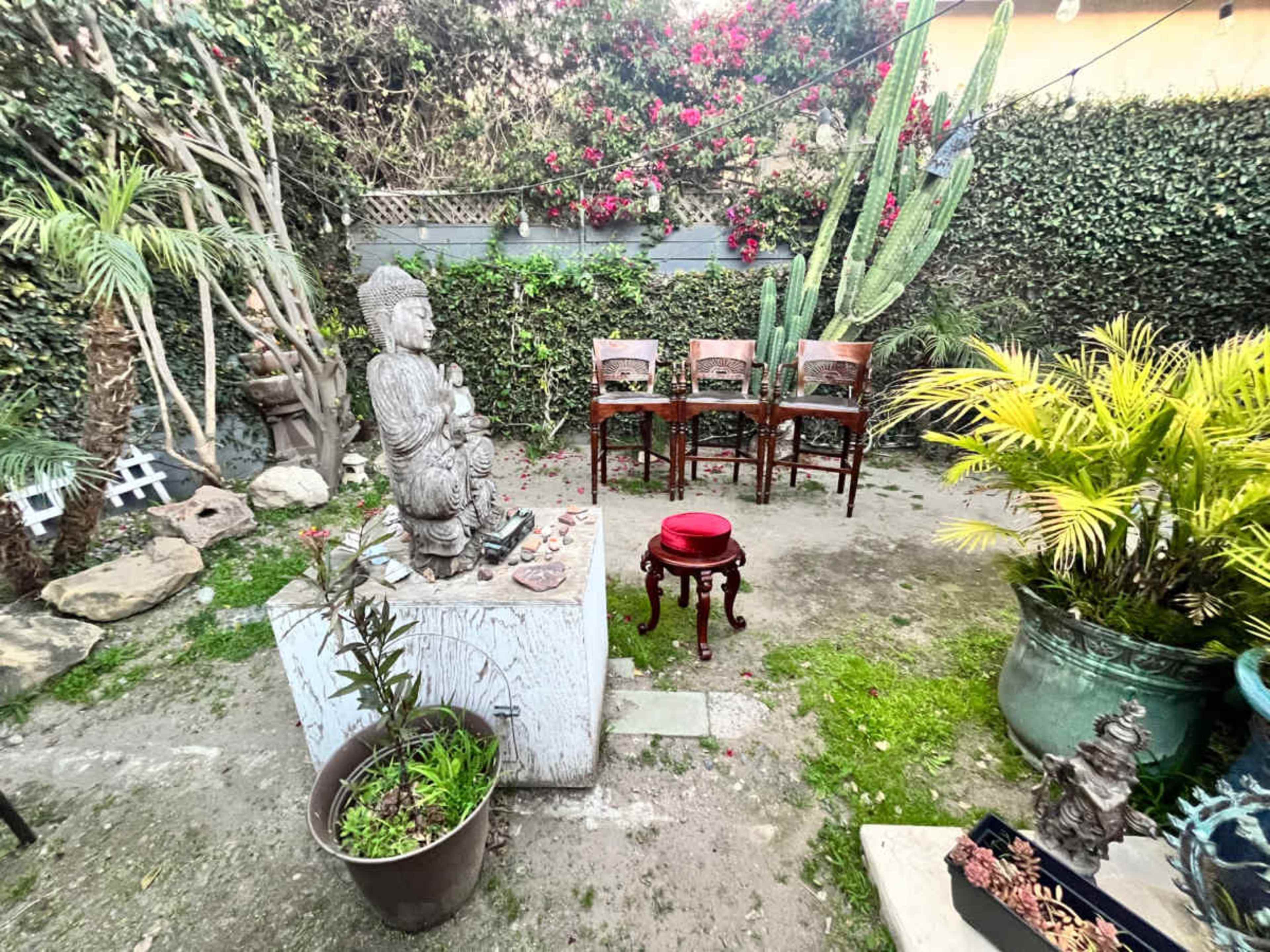 The scene features a small garden with a stone statue, several potted plants, and four wooden chairs arranged around a central red stool.