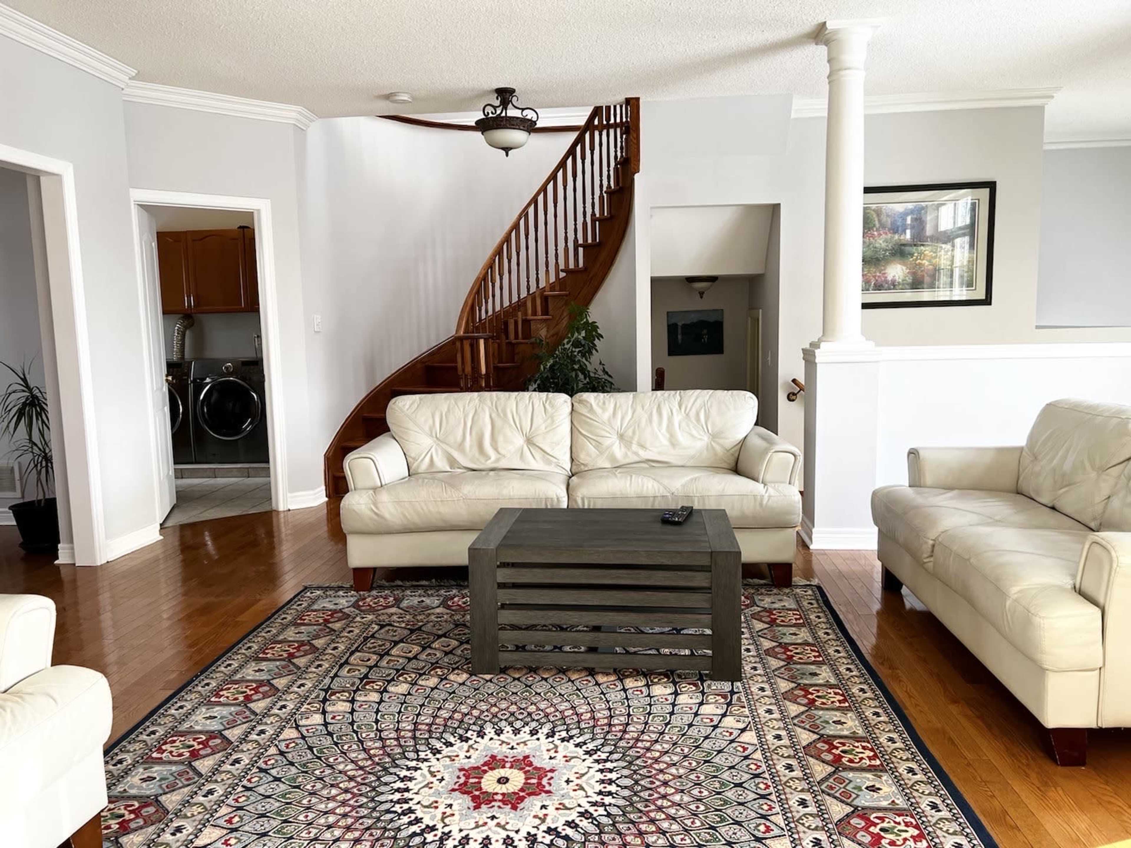 A living room with two white couches facing a wooden coffee table, a patterned area rug on hardwood flooring, and a staircase in the background.