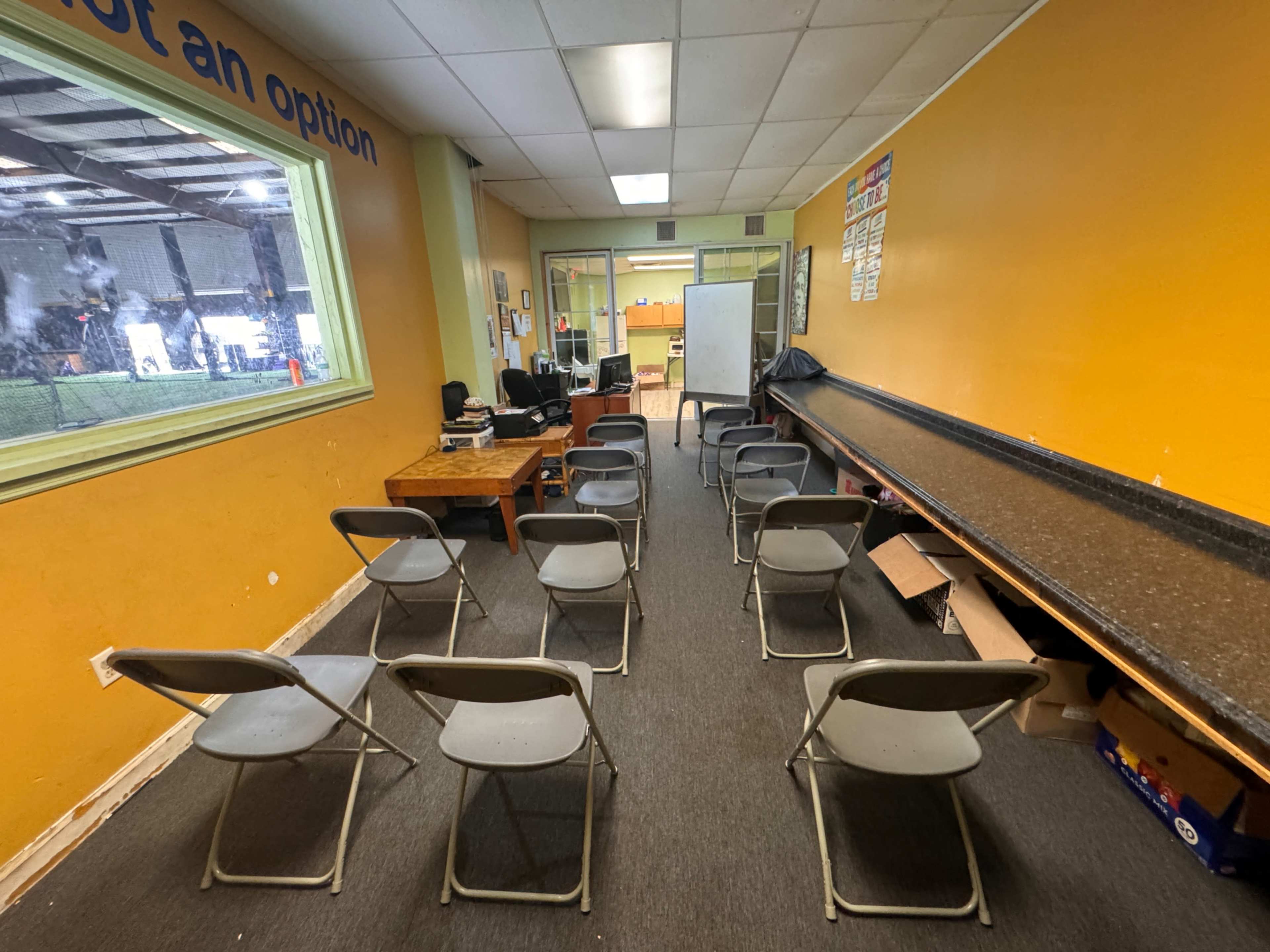 A room with a row of gray folding chairs facing a whiteboard, a desk with equipment in the corner, and a window revealing an outdoor area.