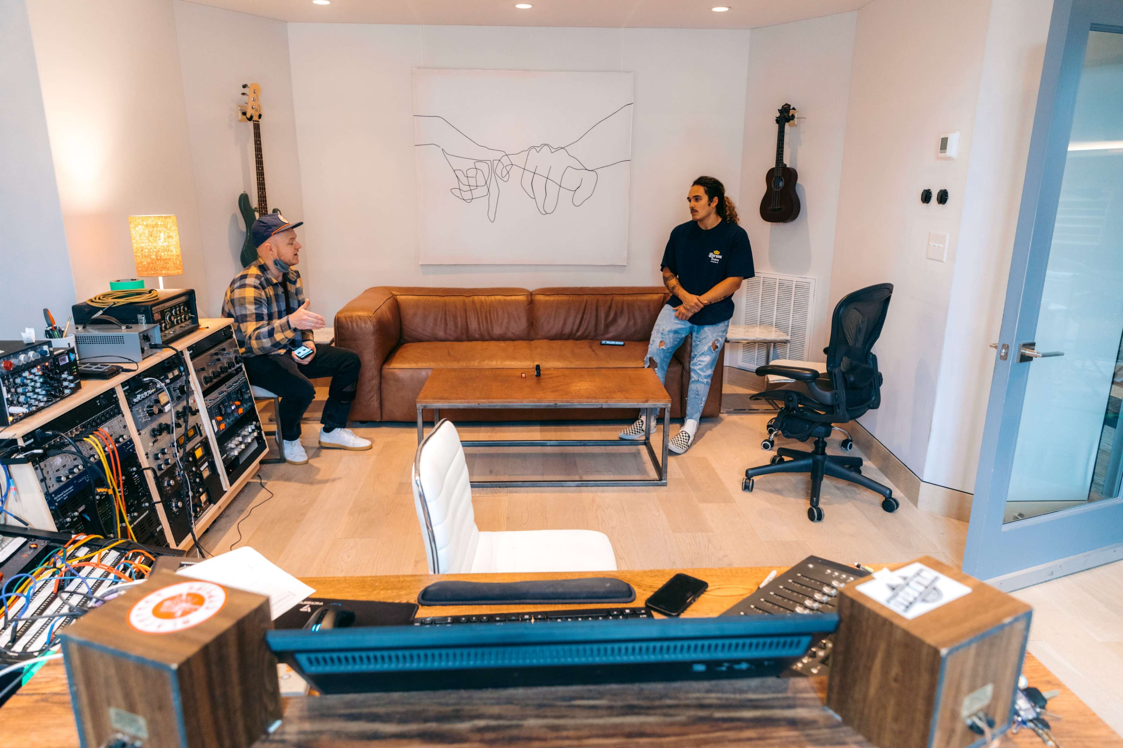 Two people converse in a modern studio space featuring a leather couch, a glass coffee table, and musical equipment.