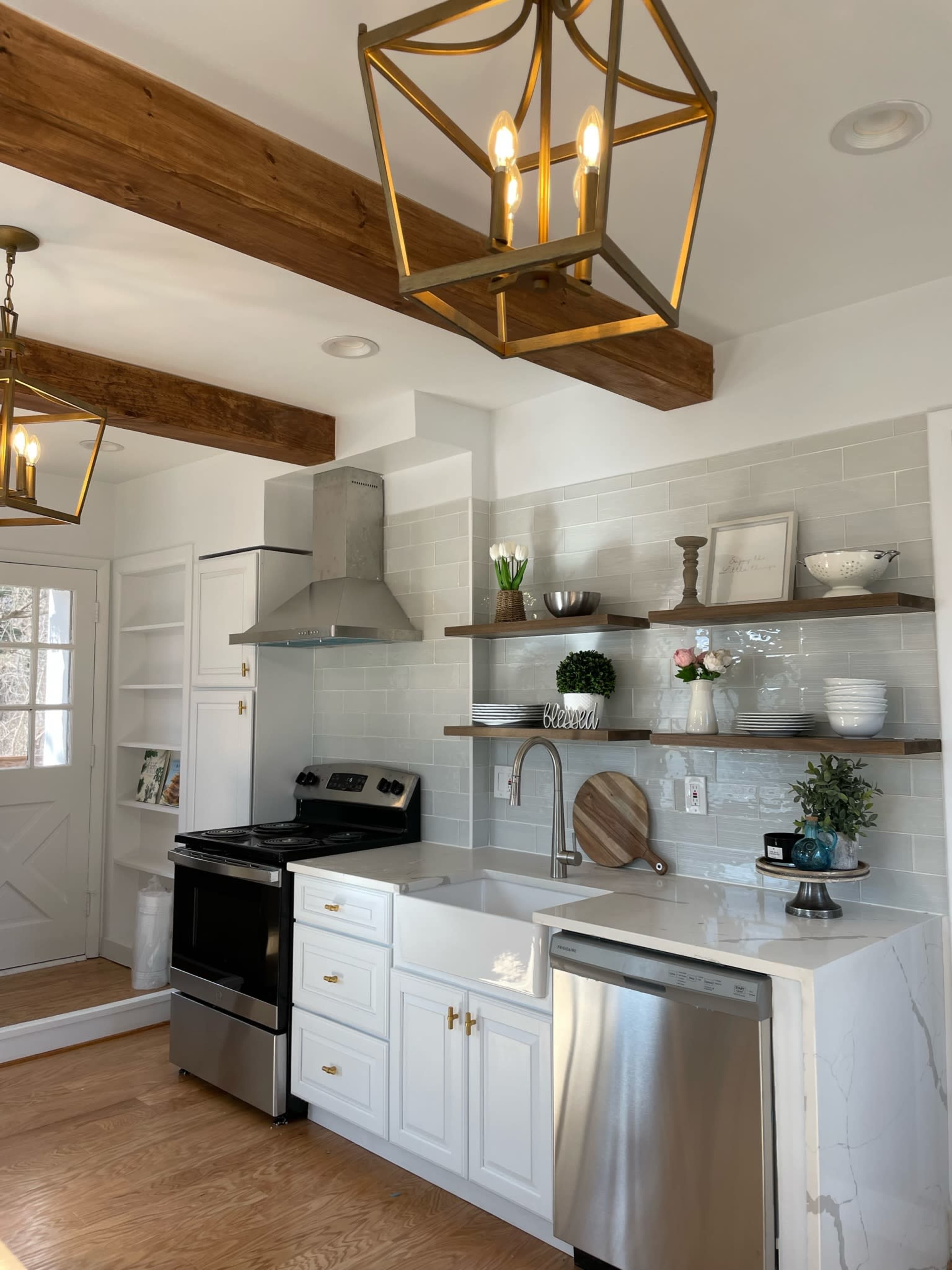 The kitchen features white cabinetry, a stainless steel stove and dishwasher, open shelving, and pendant lighting, with a door leading to another room.