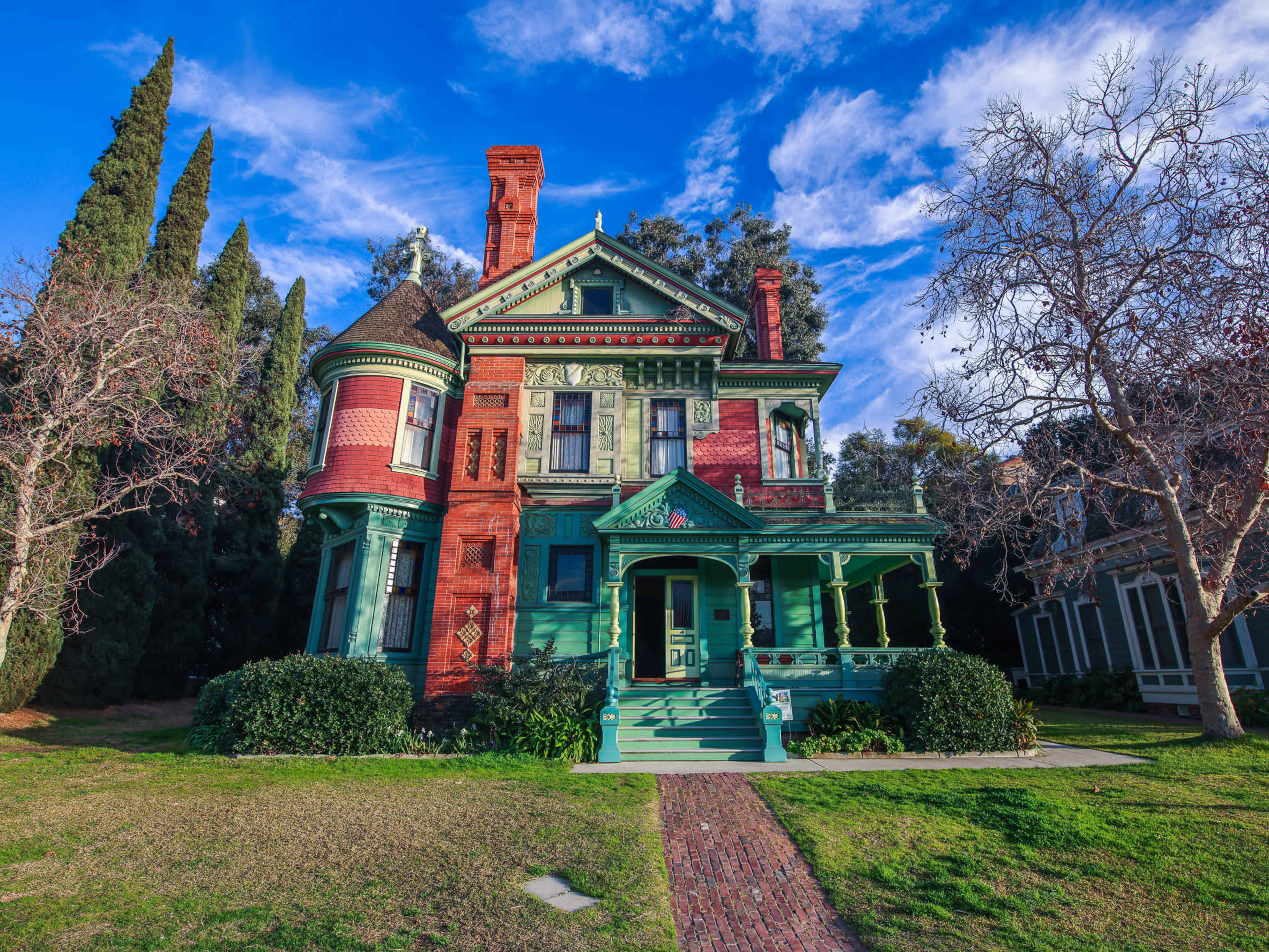 A Victorian-style house features red brick and green trim with ornate architectural details, surrounded by manicured lawn and trees.