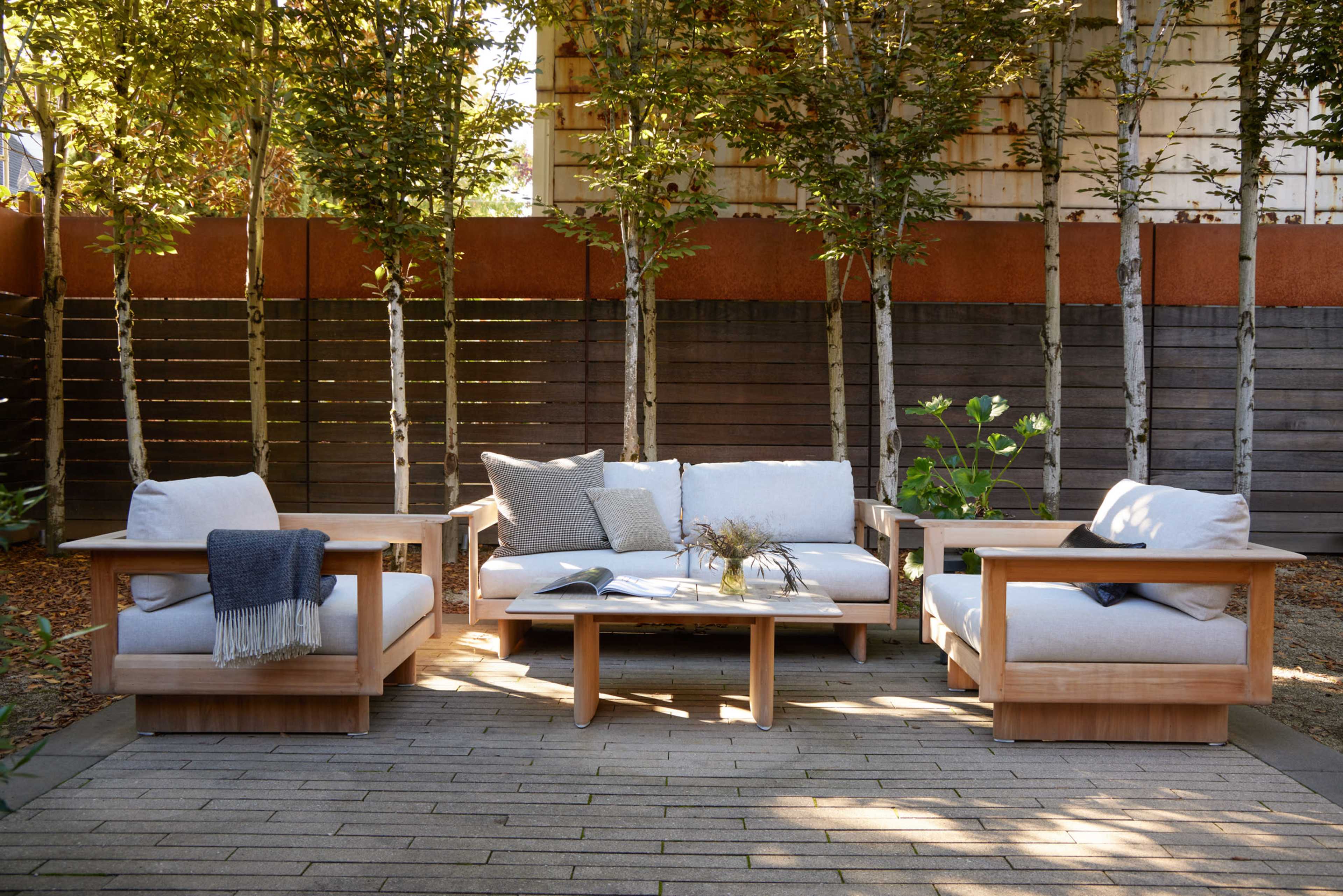 The image shows a patio area with a wooden seating arrangement consisting of two armchairs, a sofa, and a coffee table, surrounded by slender trees and a wooden fence.