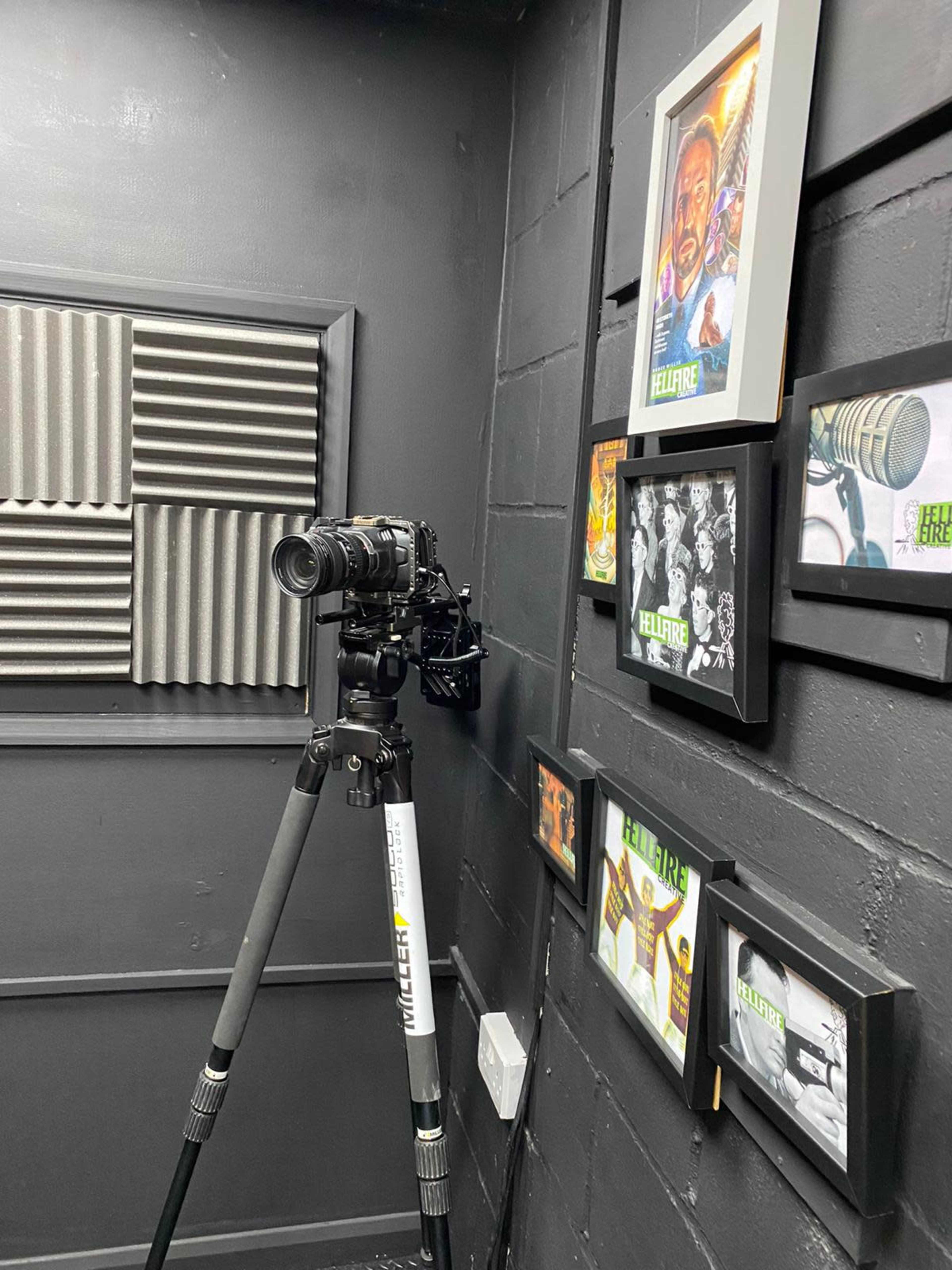 A camera on a tripod is positioned beside framed artwork on a black wall, with sound-absorbing panels in the background.
