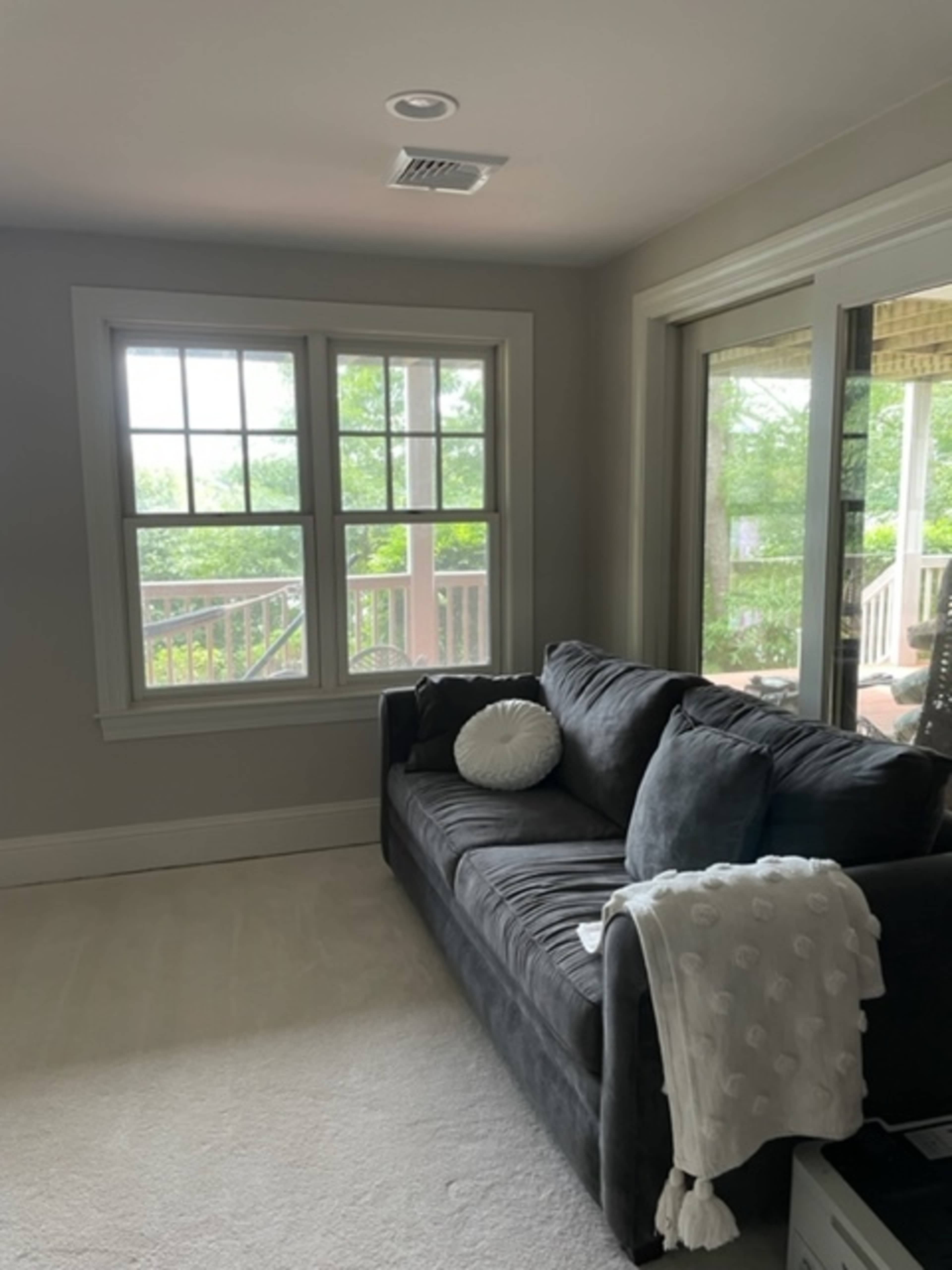 A gray sofa with a white throw pillow is positioned against a wall with two large windows overlooking a porch.