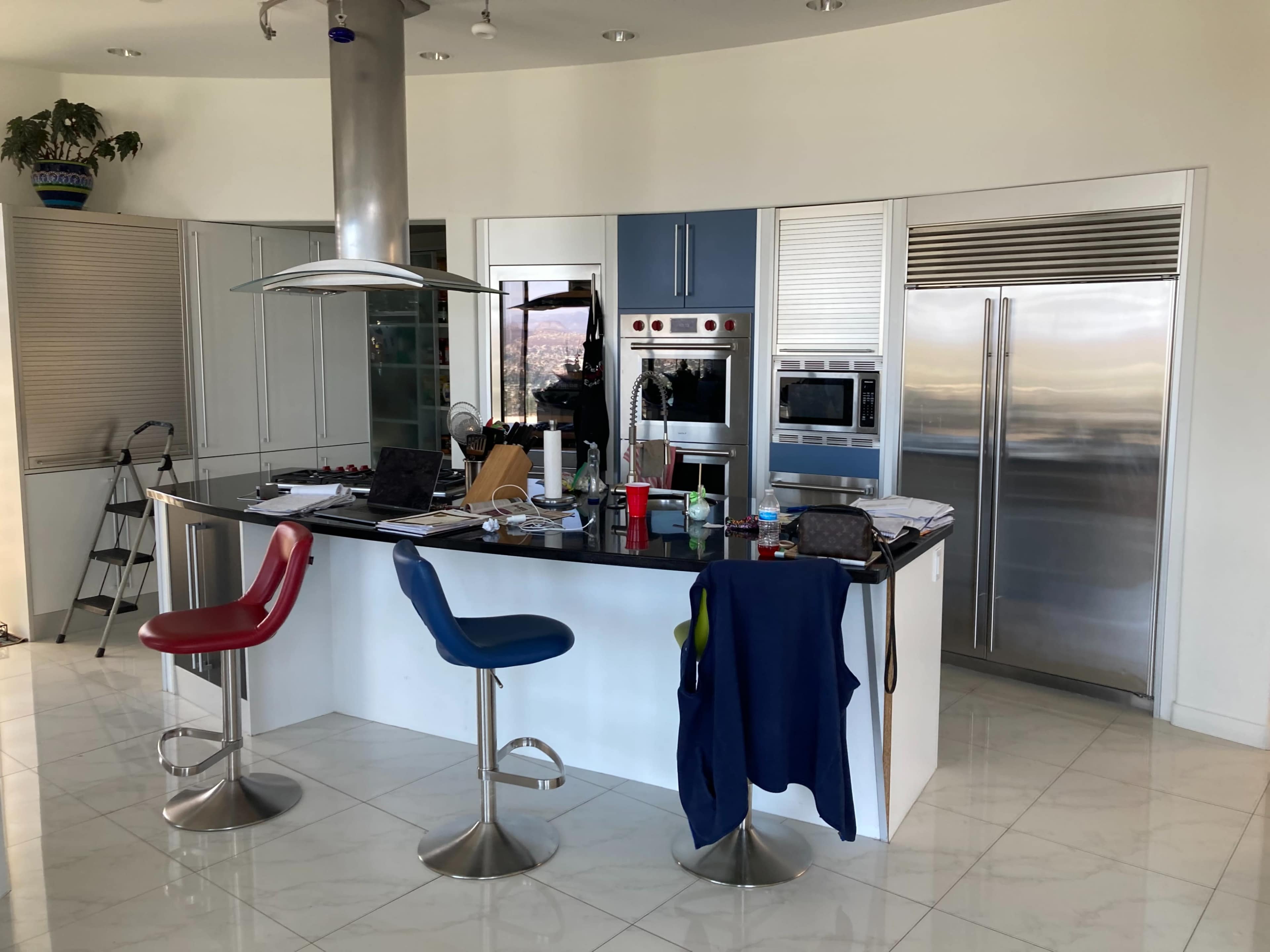 A modern kitchen with a central island, featuring bar stools, appliances, and various items scattered on the surfaces.