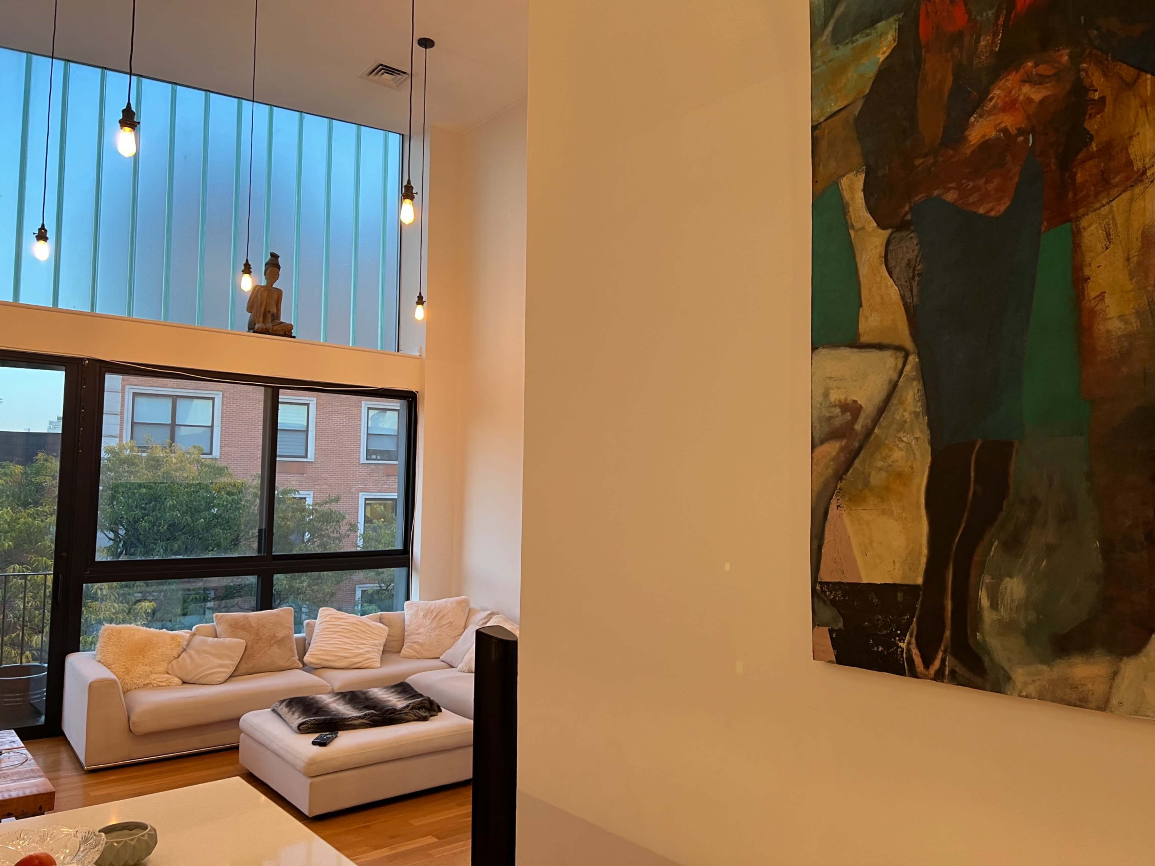 A modern living room featuring a large white sofa, large windows with natural light, and a colorful abstract painting on the wall.