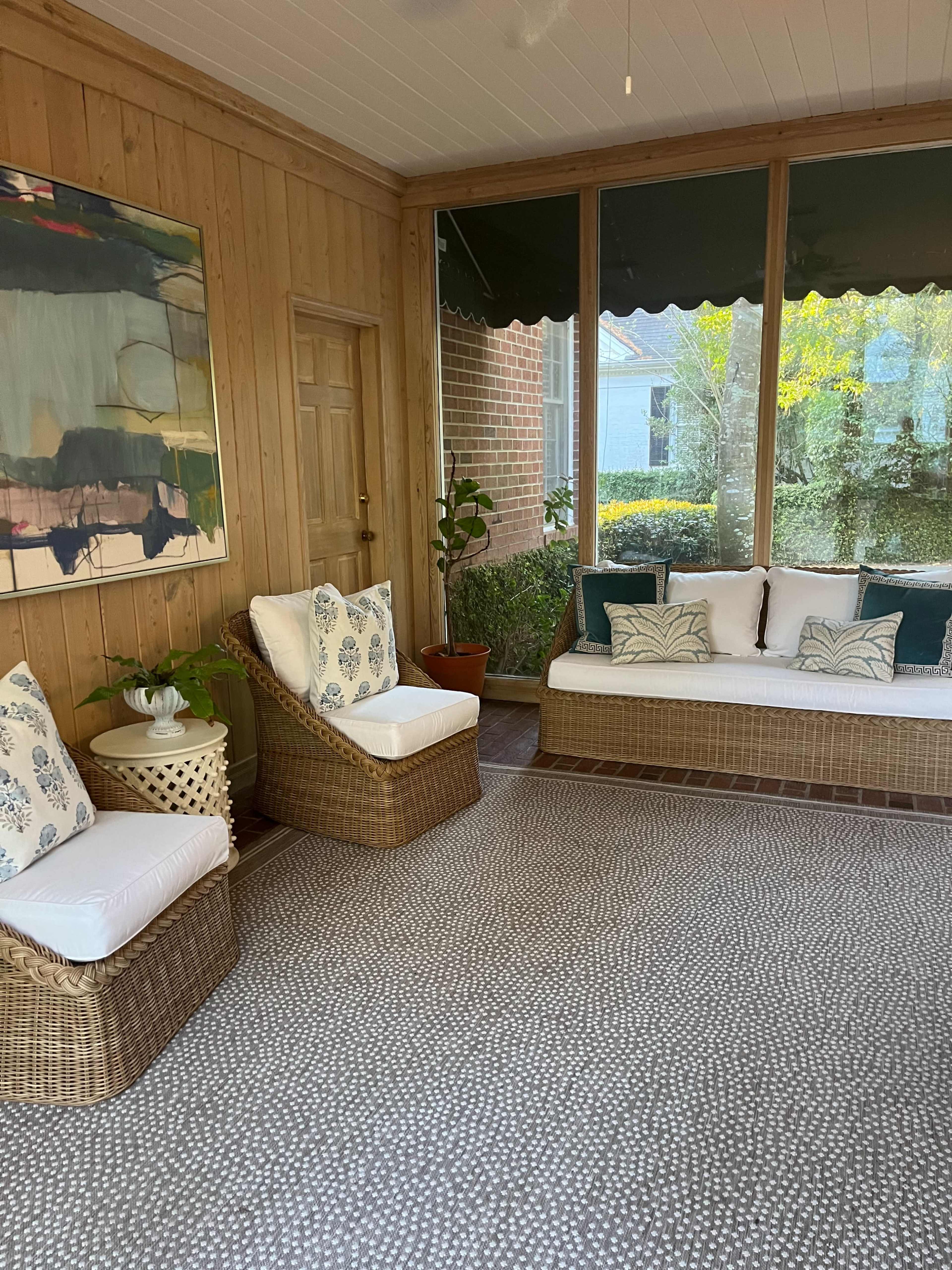 The image shows a sunroom with wicker seating, a large abstract painting on the wall, and a patterned rug.