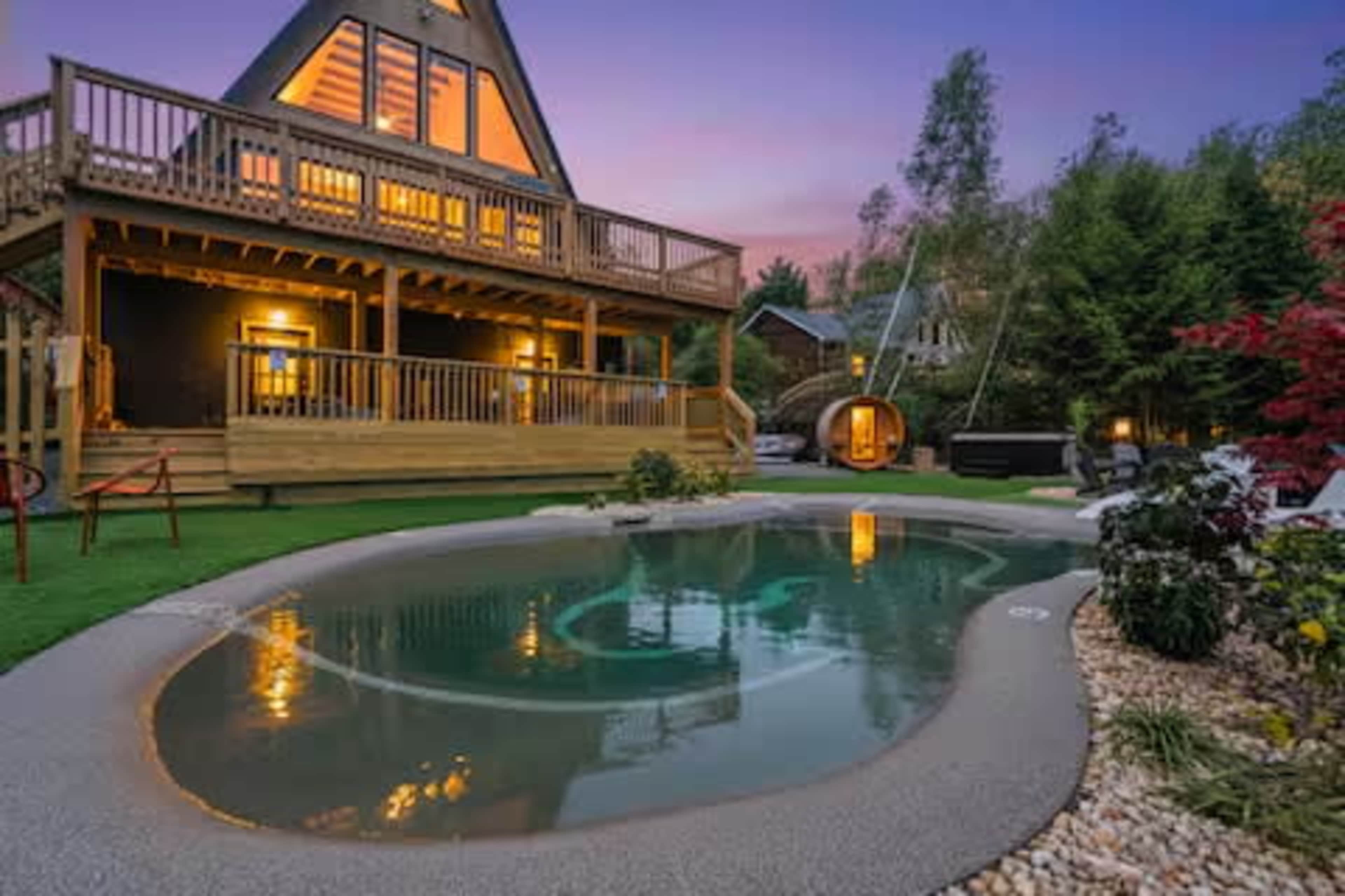 A modern wooden chalet with large windows overlooks a landscaped backyard featuring a small pool and surrounding greenery at twilight.