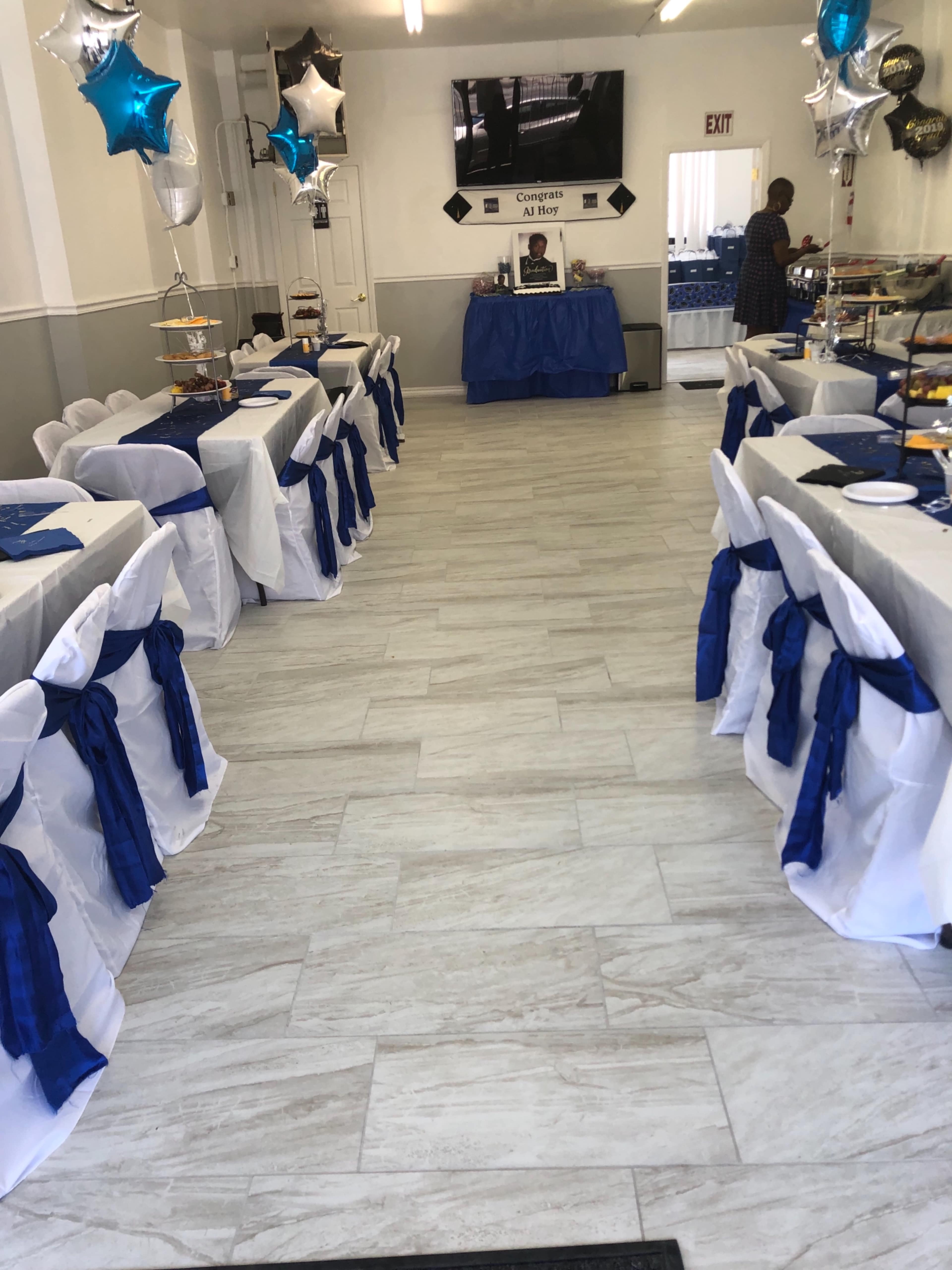 The image shows a dining area with neatly arranged tables covered in white tablecloths and blue sashes, along with balloons and a large screen display at the front.