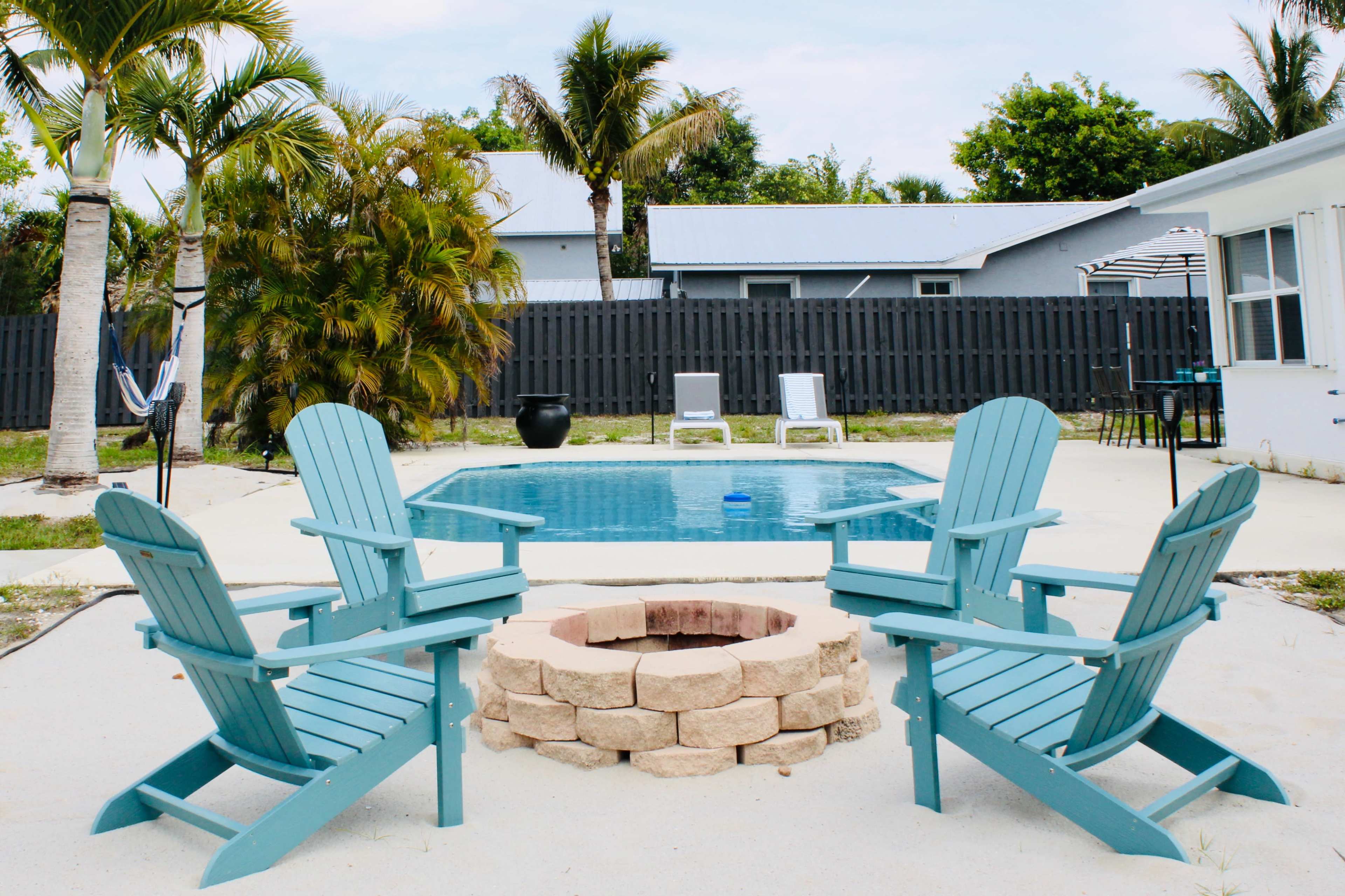 A backyard with a pool surrounded by four blue Adirondack chairs arranged around a stone fire pit, palm trees, and a gray wooden fence in the background.