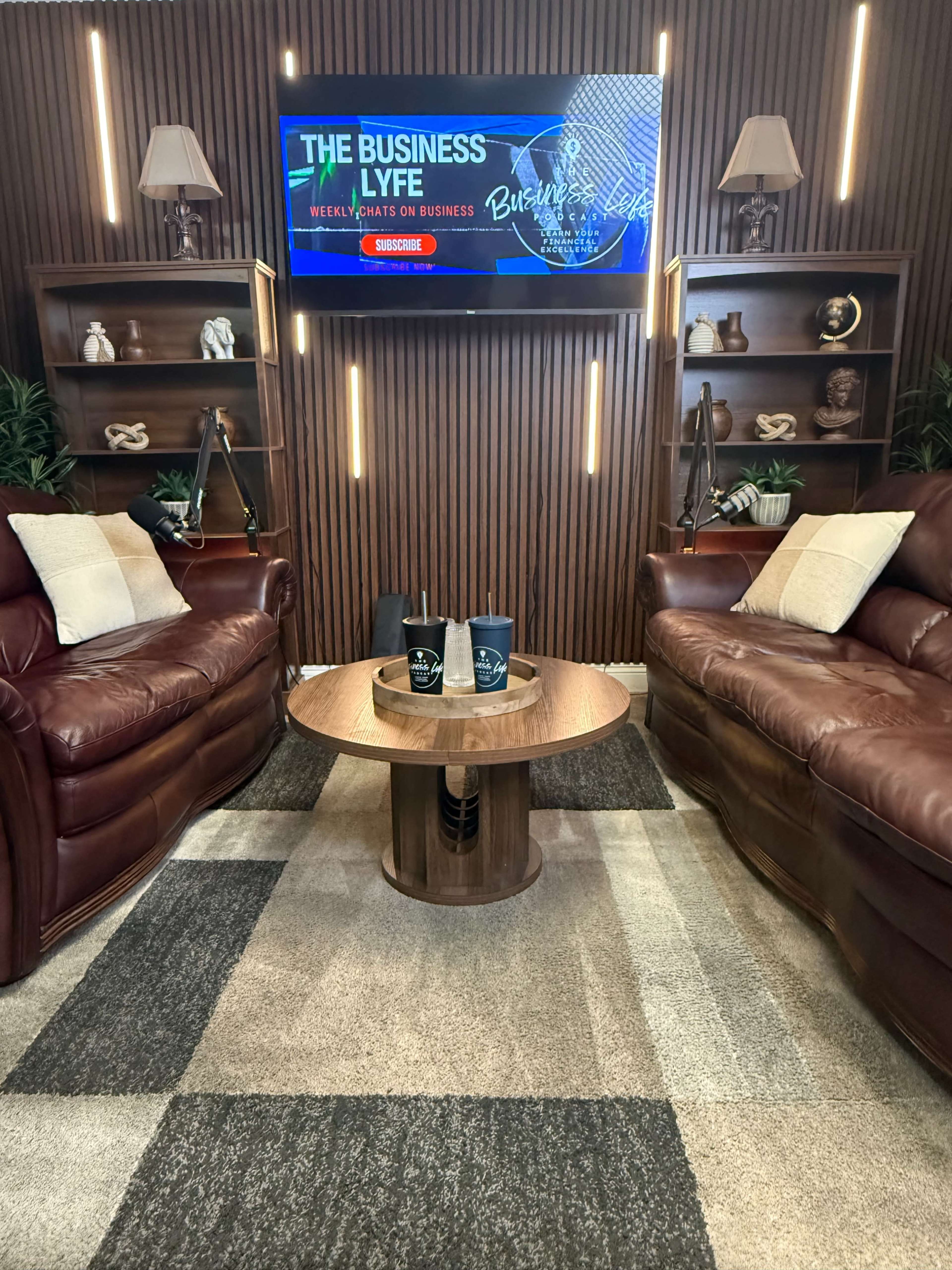 The image shows a cozy indoor setting with two leather sofas facing a wooden table, framed by shelves and a large screen displaying "The Business Lyfe" and a "Subscribe" button.