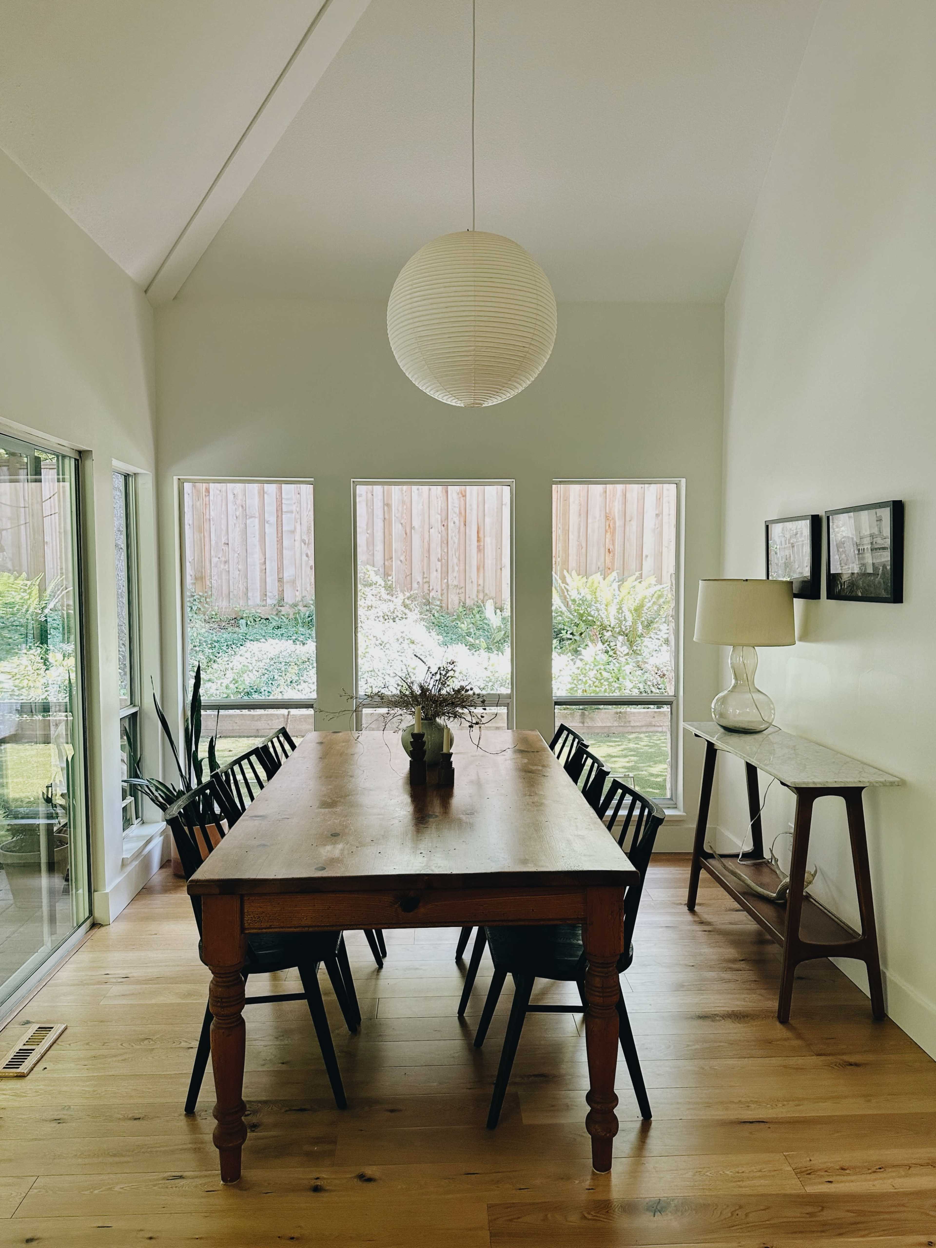 A wooden dining table with black chairs is centered in a bright room that features large windows overlooking a garden.