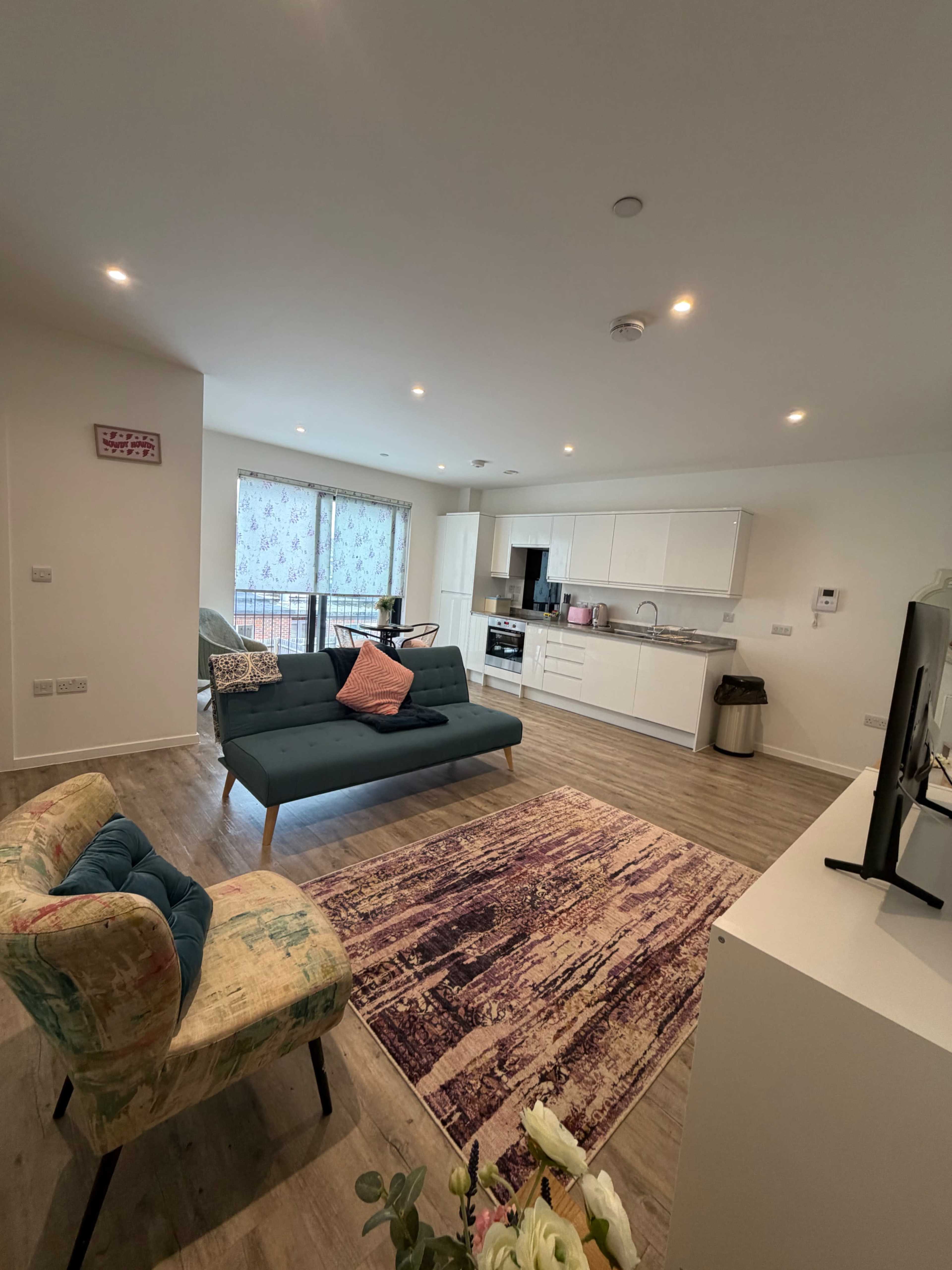 The image shows a modern, open-plan living space with a teal sofa, a colorful armchair, a kitchen area, and large windows letting in natural light.